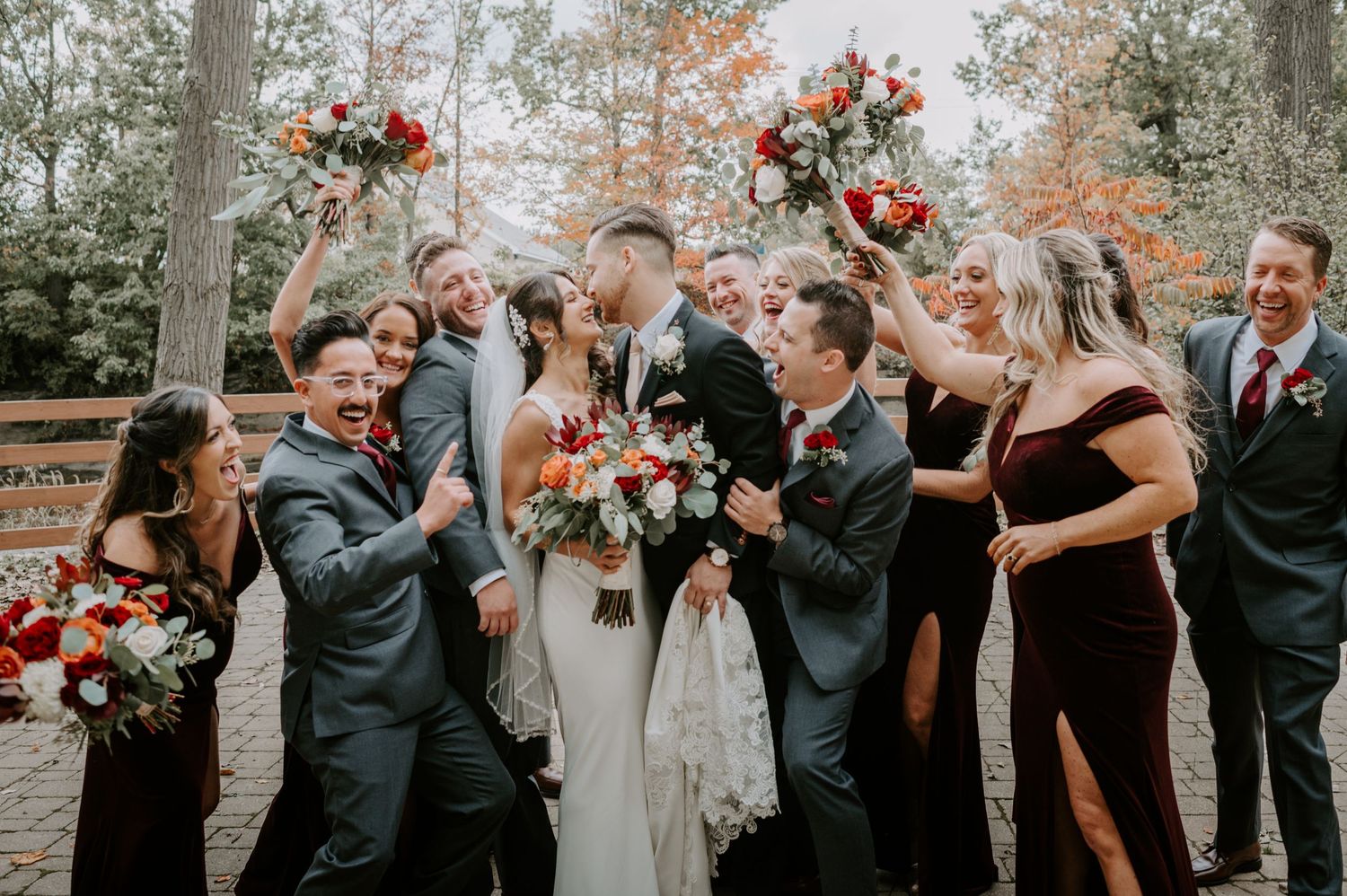 Bride and groom kiss while surrounded by a cheering wedding party in an outdoor fall setting.