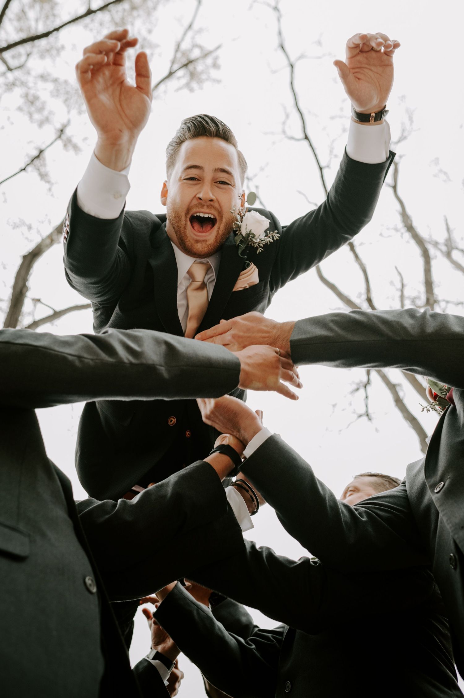 Groomsmen lift a joyful groom into the air as he celebrates with arms raised at his wedding.