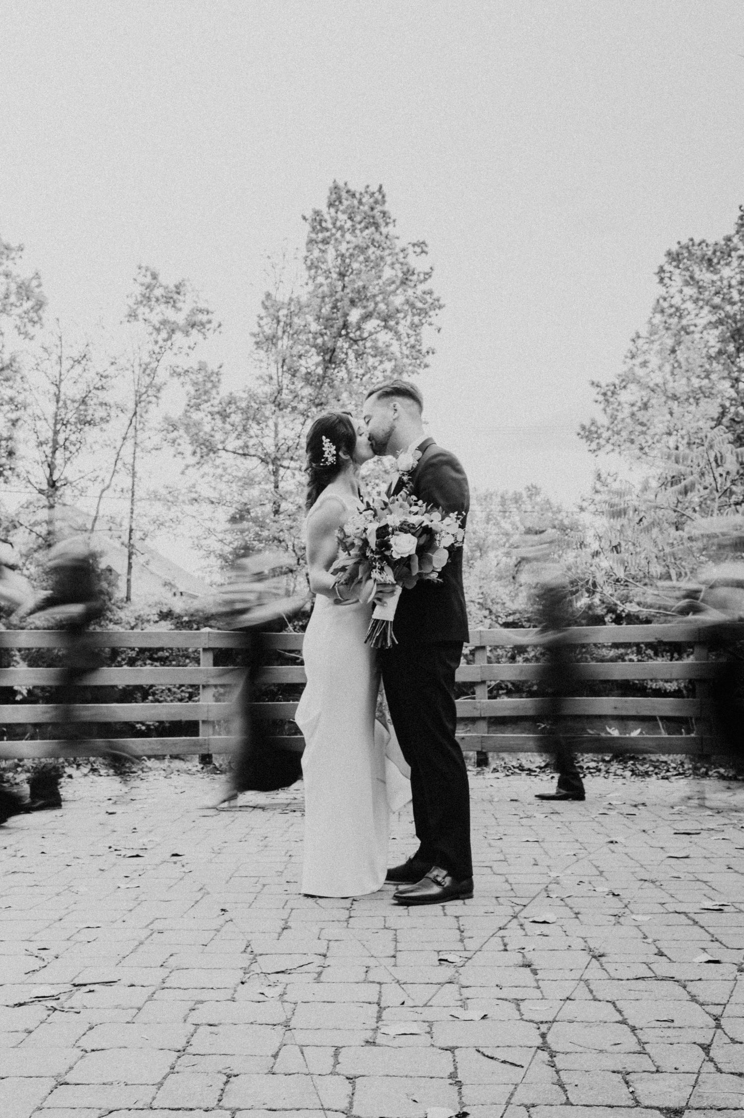 Black and white photo of bride and groom kissing on a cobblestone patio surrounded by trees.