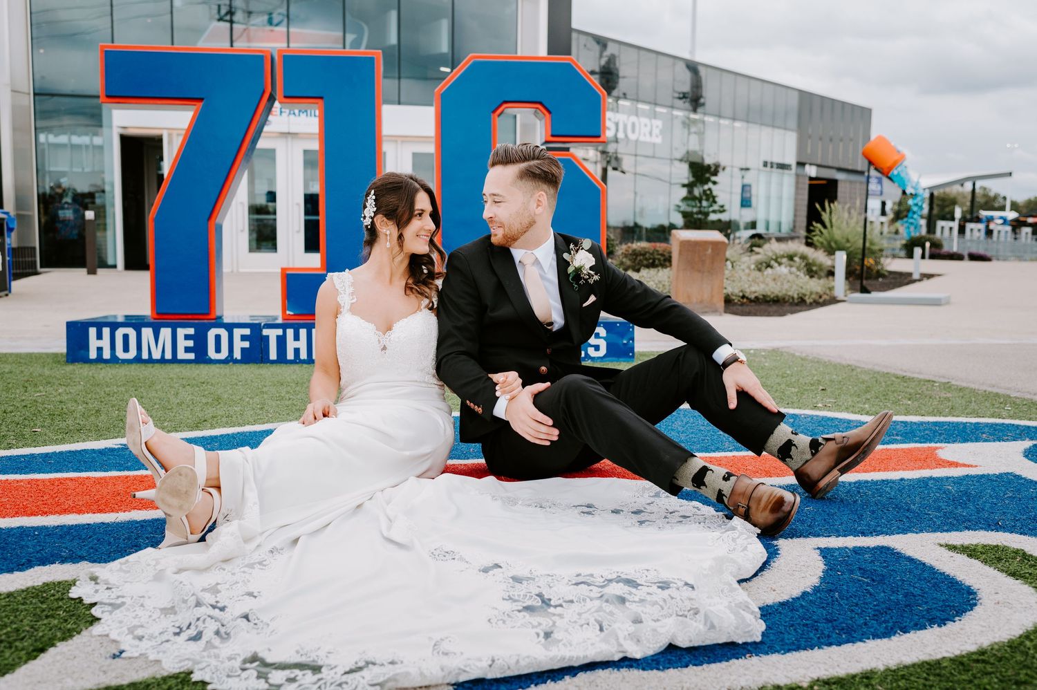 Bride and groom sit back-to-back on a football field logo in front of a stadium 716 sign.