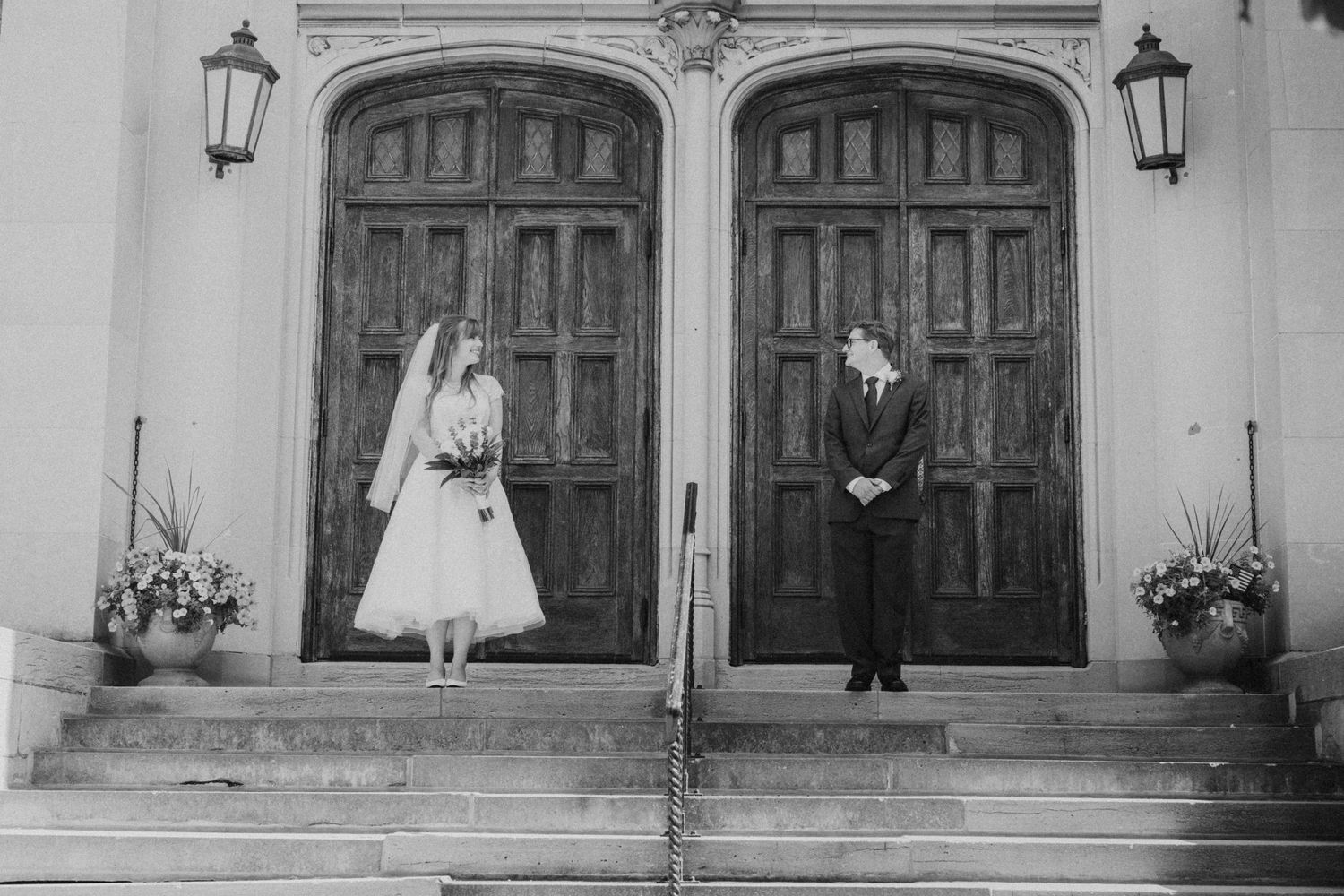 Black and white photo of bride and groom standing apart on church steps before grand wooden doors.