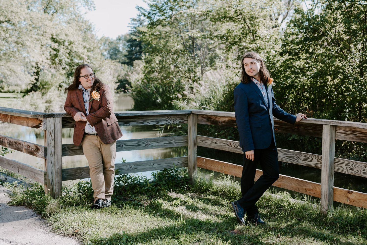 Two women in blazers leaning against a wooden fence bridge surrounded by green trees.