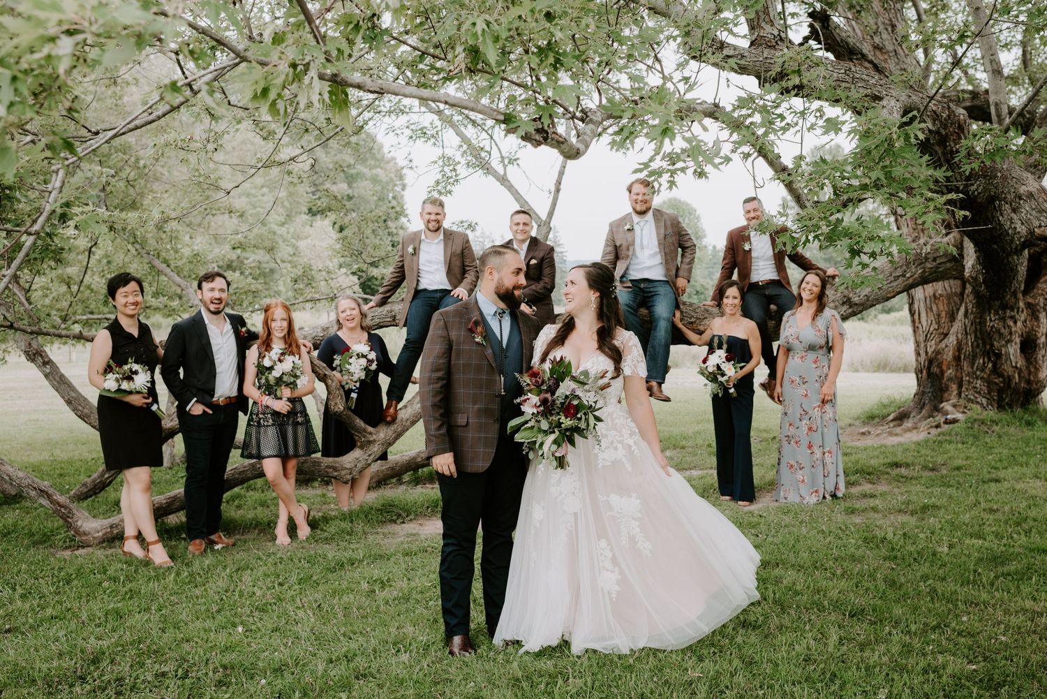 Wedding party posing with bride and groom under a large tree in an outdoor rustic setting.