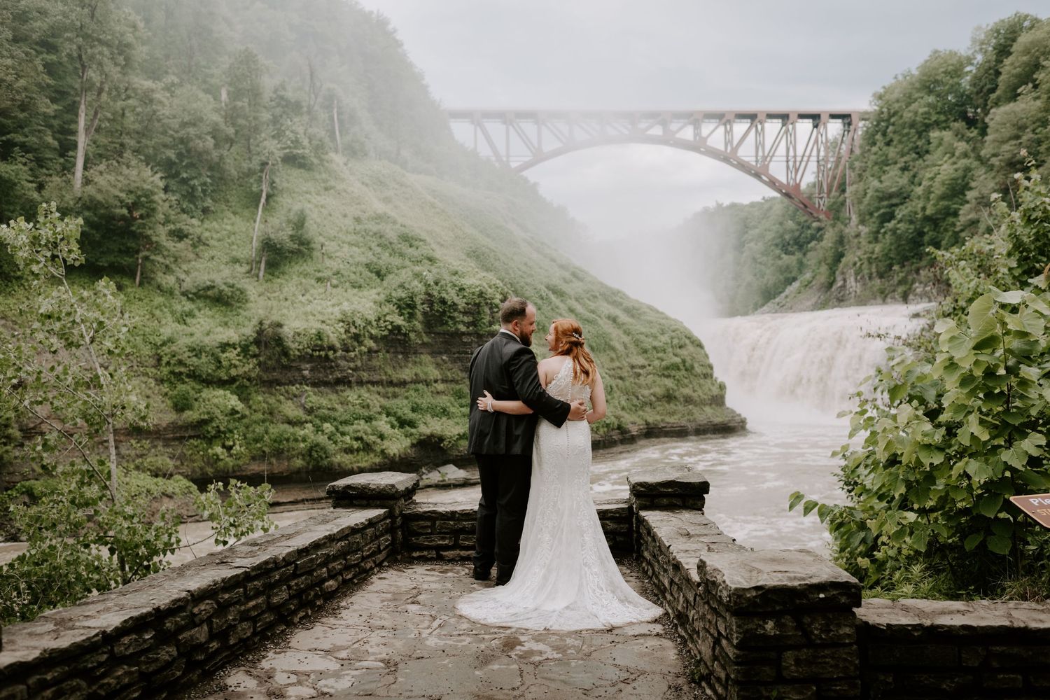Bride and groom sharing a romantic moment near a stunning waterfall with a stone bridge backdrop.