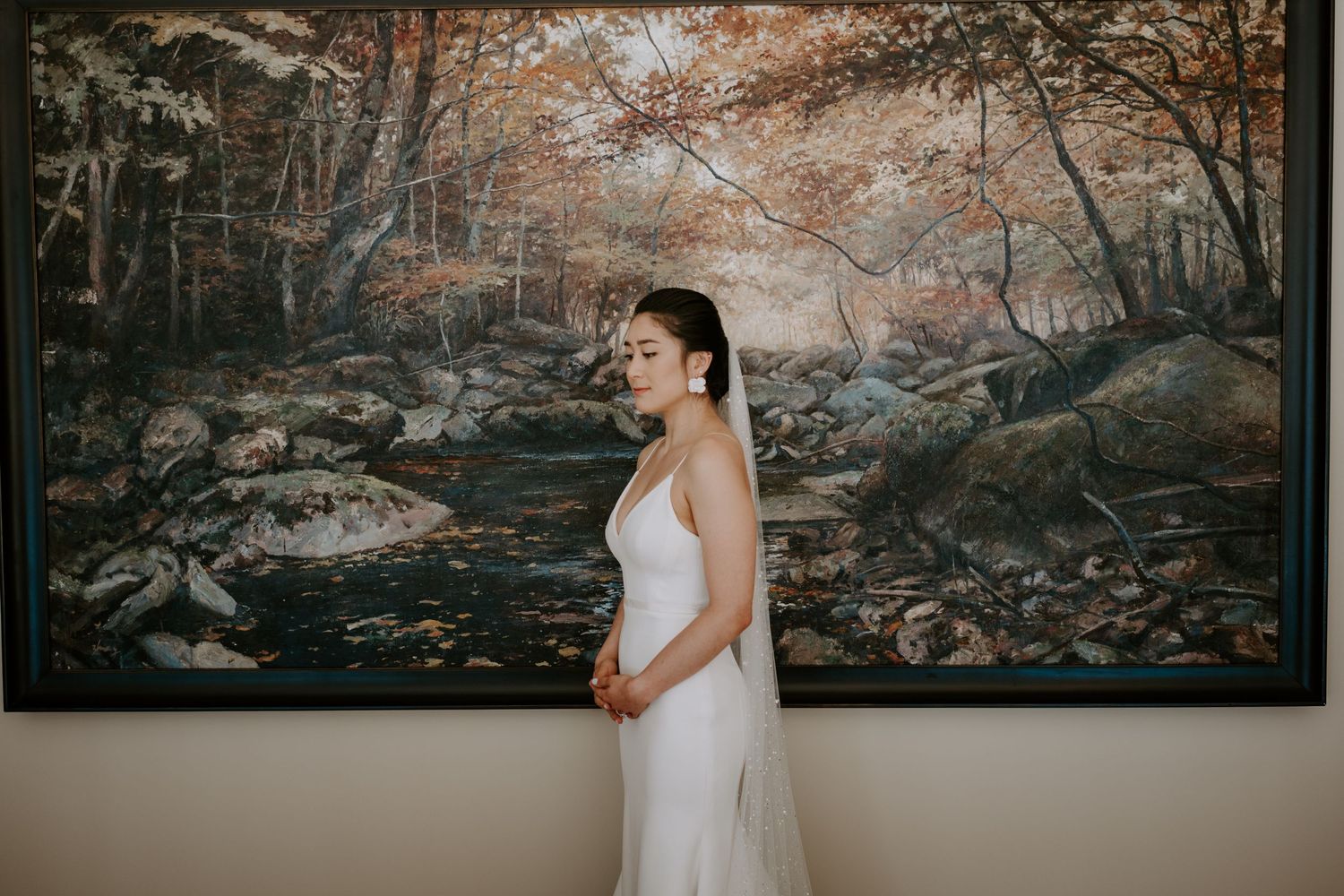 Elegant bride in a white gown standing in profile before a large nature-inspired painting indoors.