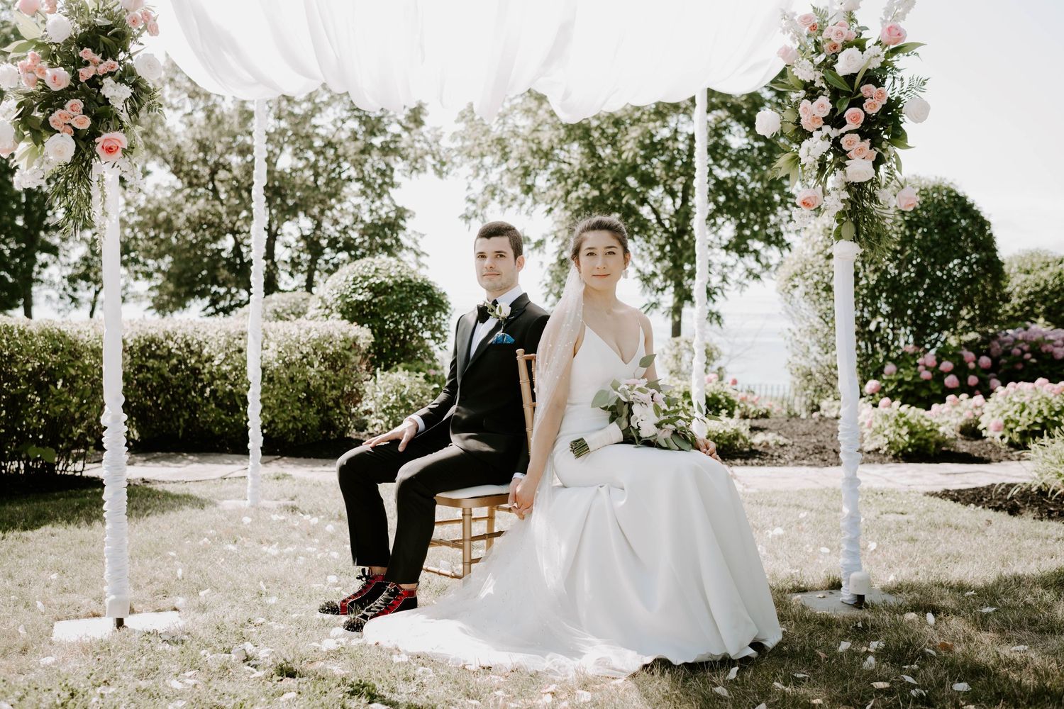 Bride and groom sitting back-to-back under a floral wedding arch in a lush outdoor garden ceremony.