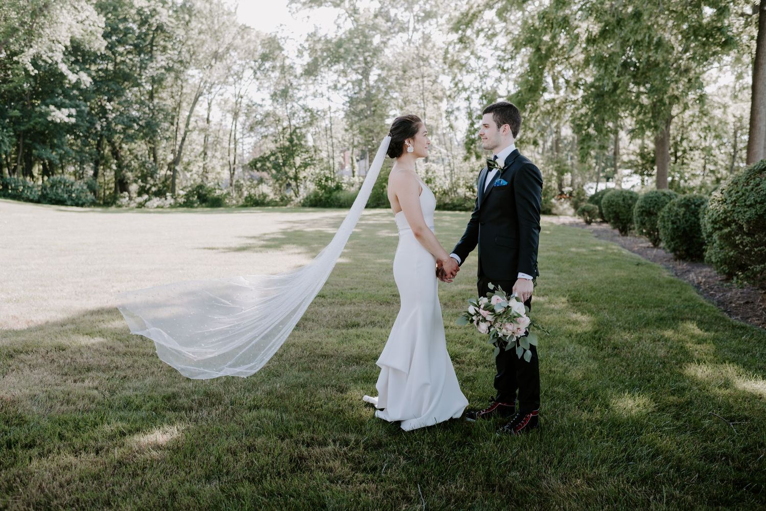 Bride and groom holding hands on a sunlit lawn with her veil flowing gracefully in the breeze.