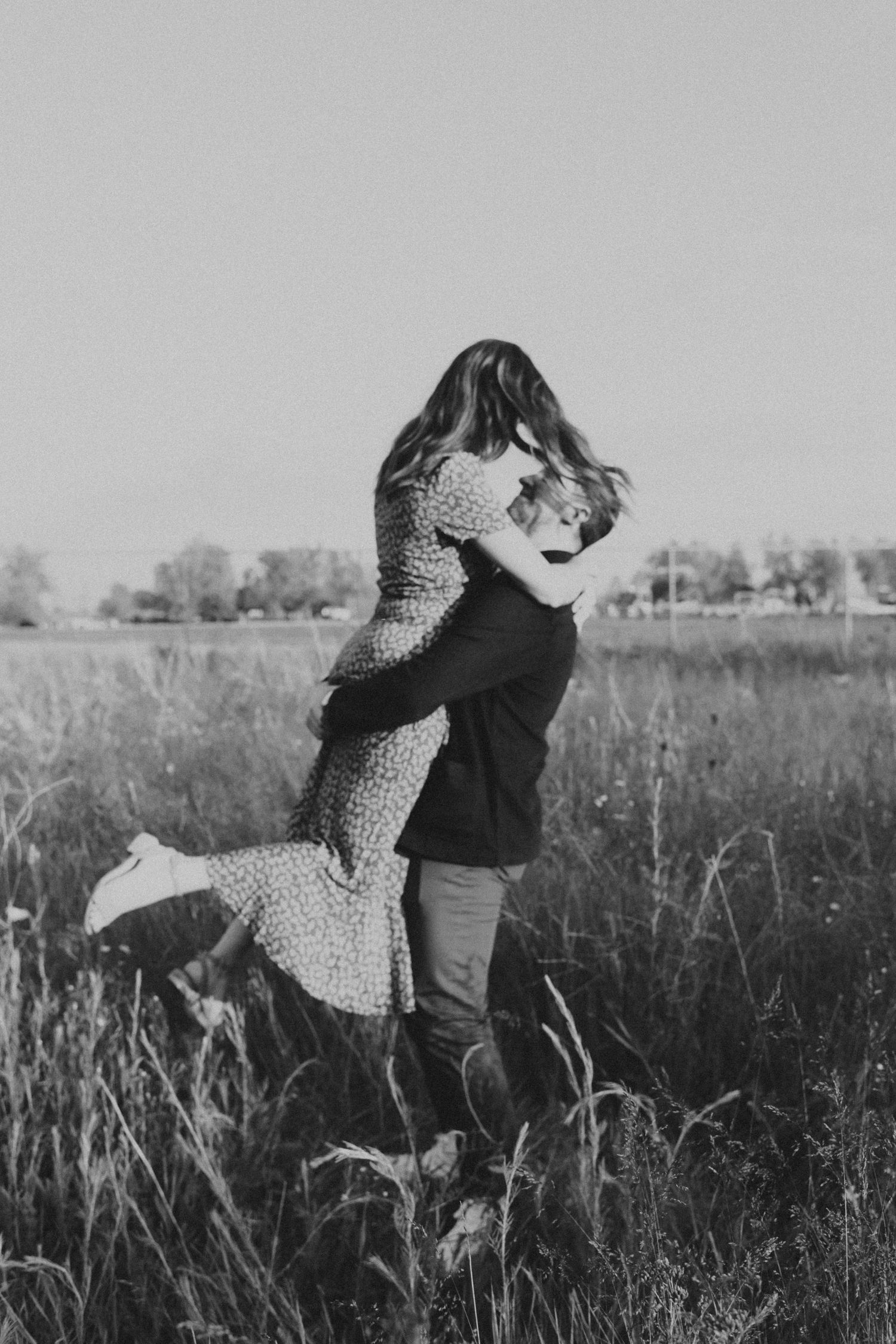Black and white photo of couple embracing in tall grass field, woman lifted off the ground.