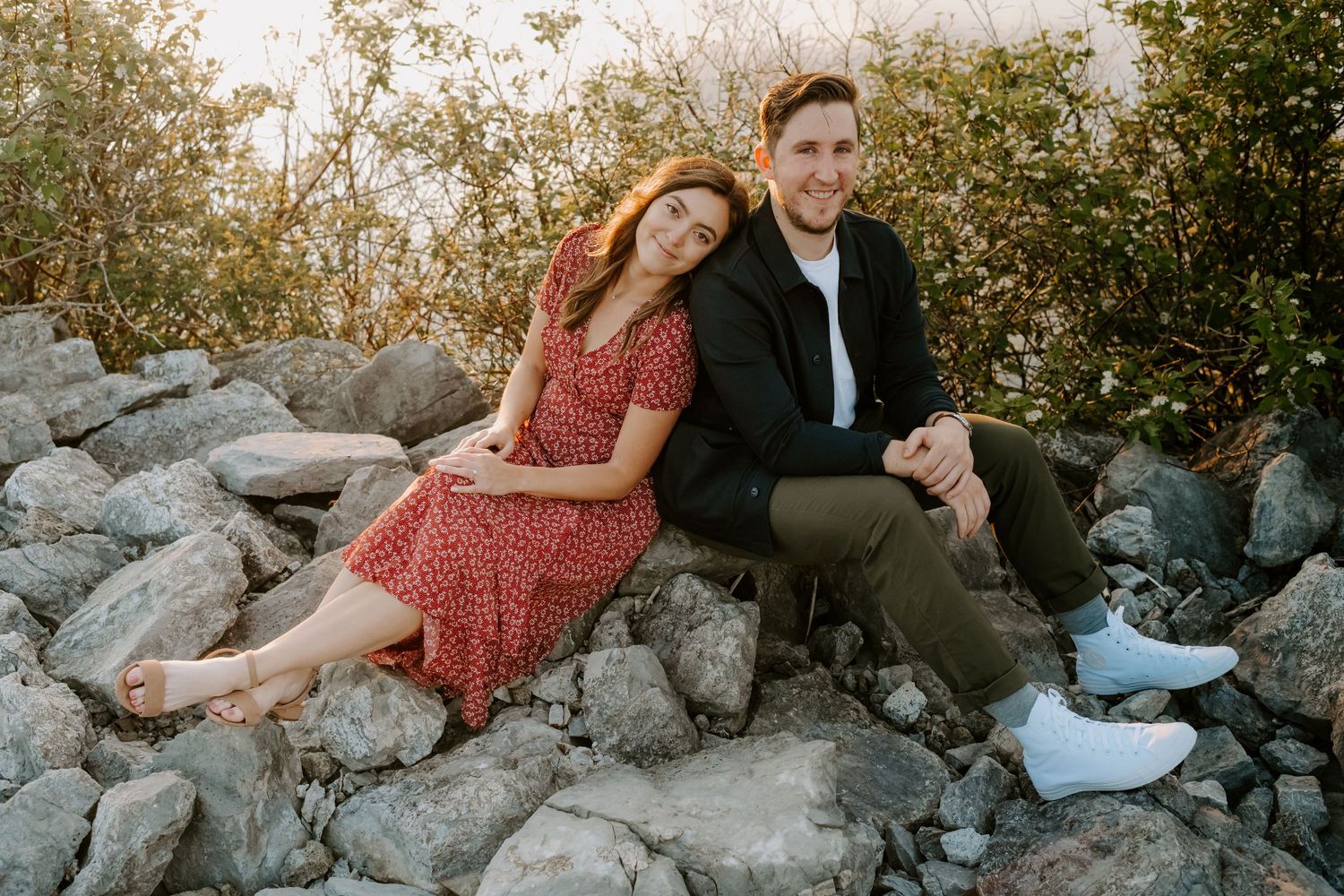 Couple sitting on rocky terrain, woman in red floral dress leaning on man in black jacket.