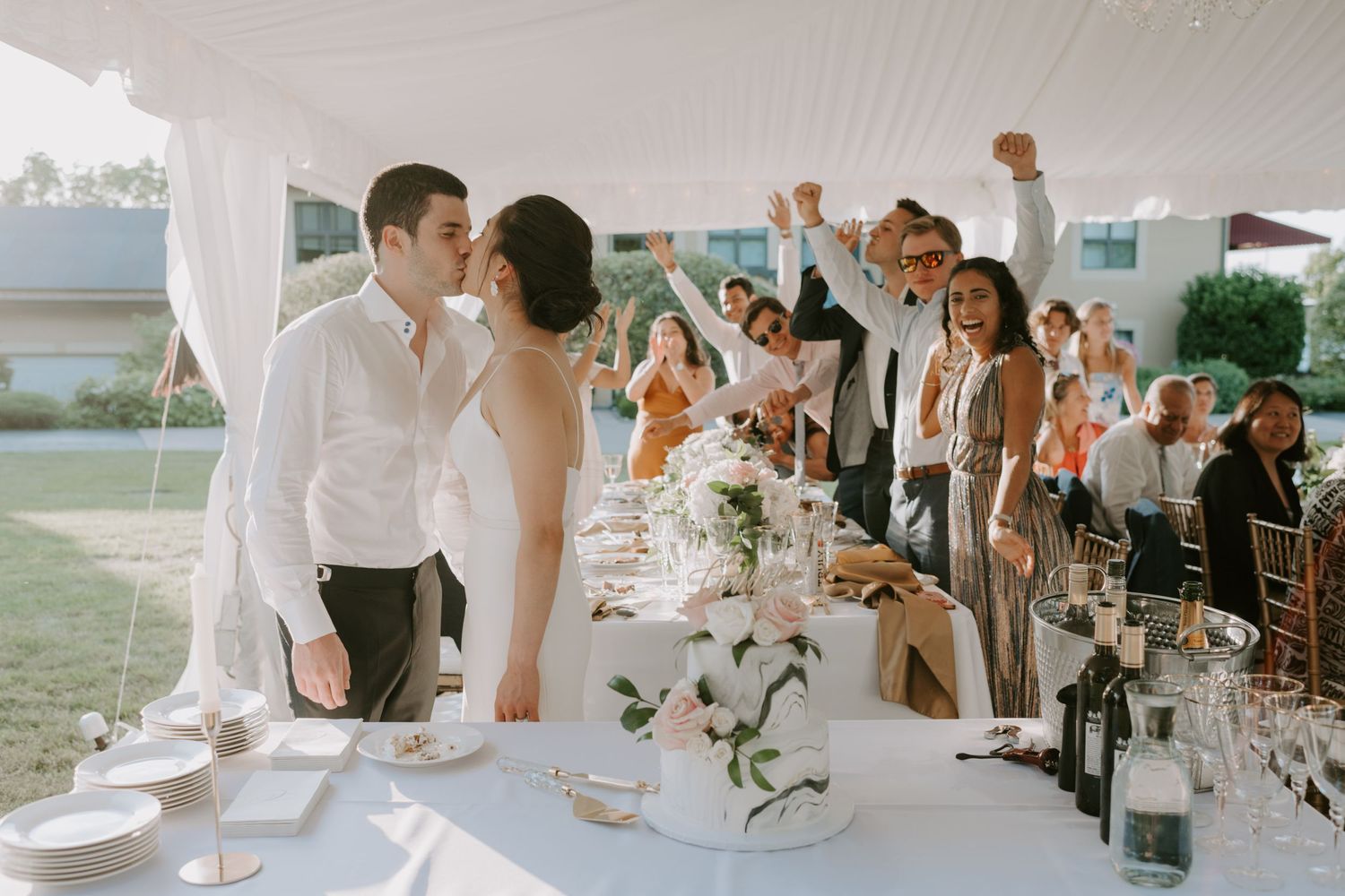 Bride and groom share a kiss at outdoor wedding reception while guests celebrate with raised arms.