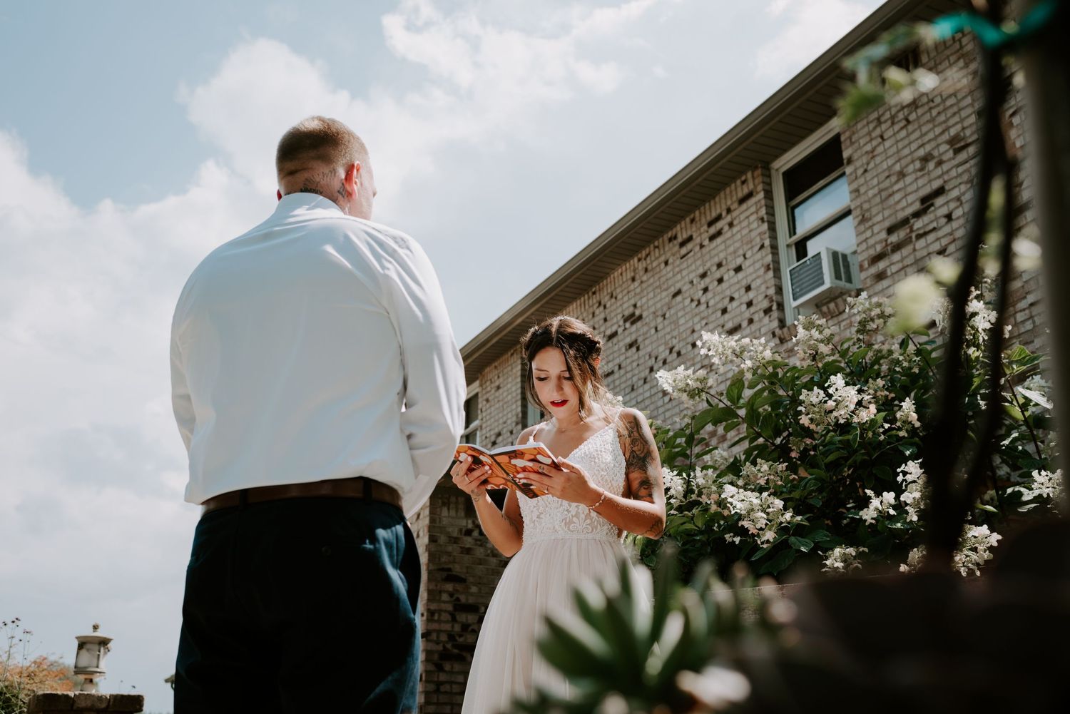 Smiling bride in white dress reads a book outside a brick building surrounded by white flowers.