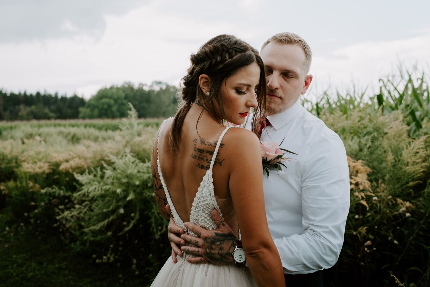 Bride with tattoos in backless gown embraced by groom in white shirt in a lush green field.