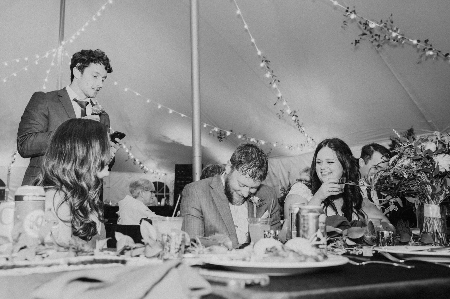Black and white photo of wedding guests laughing during a heartfelt speech at a tented reception.