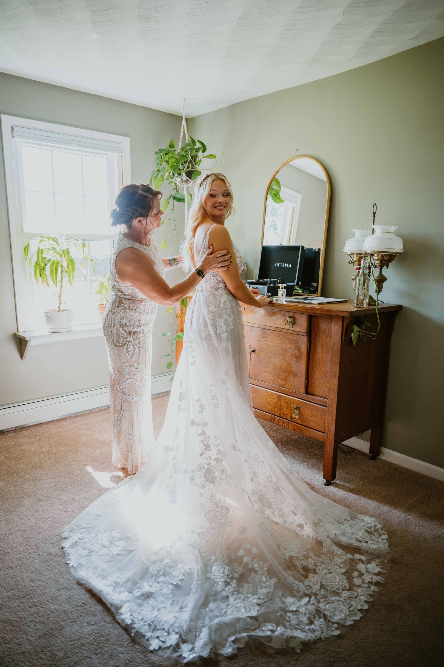 Mother helping bride into stunning lace wedding gown with long train in softly lit bedroom.