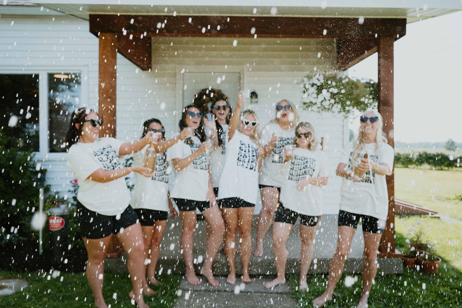Bridal party in matching white tees celebrates with champagne spray on porch before wedding.