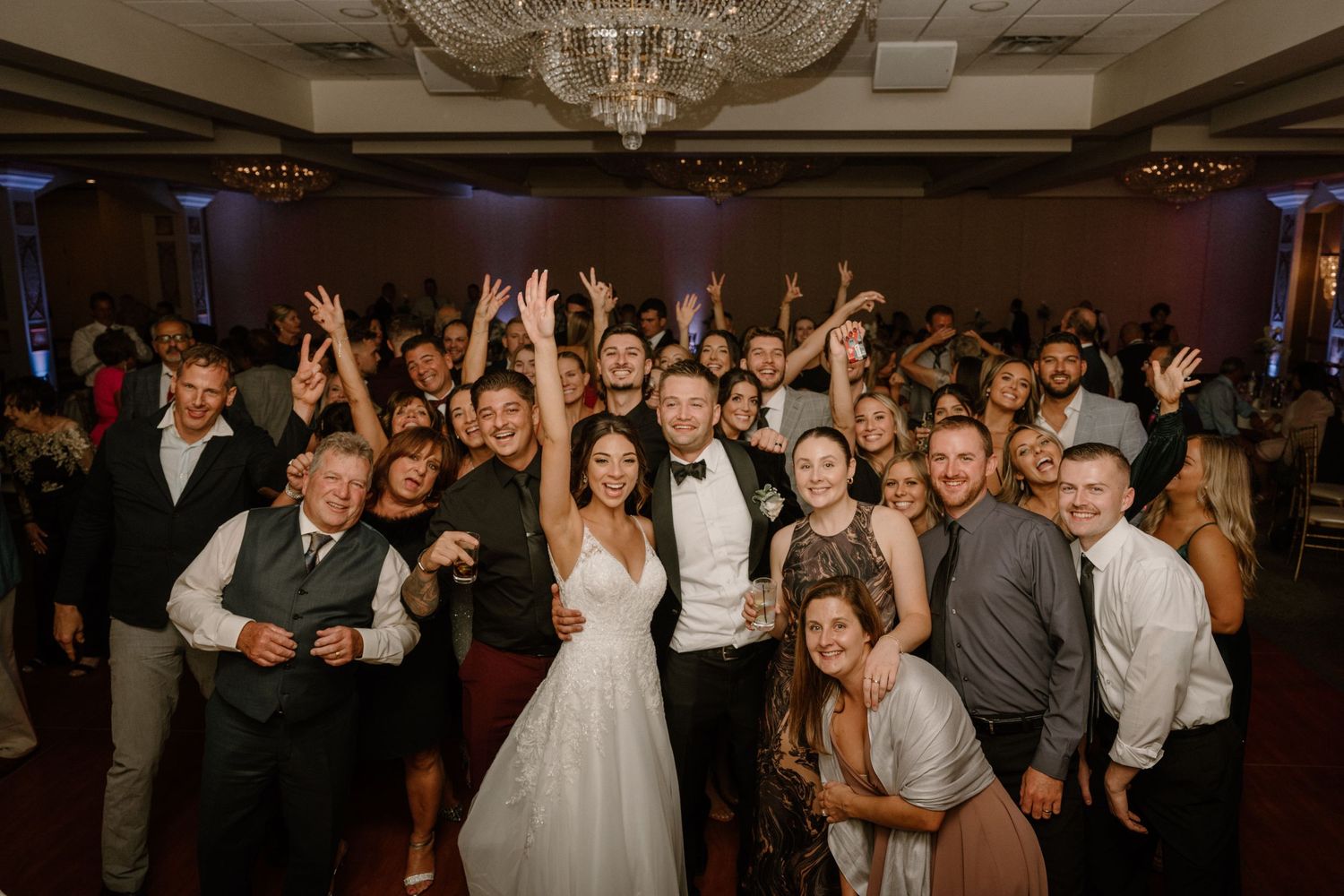Joyful wedding reception group photo with bride and groom surrounded by celebrating guests under a chandelier.