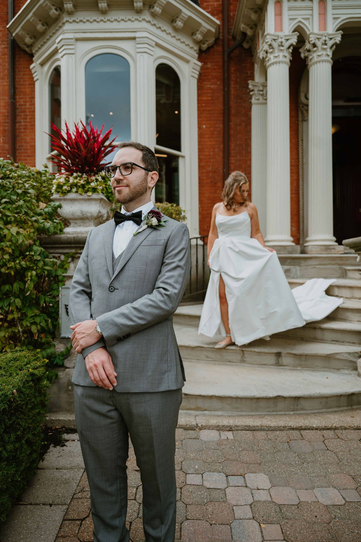 Groom in gray suit stands outside historic brick building as bride descends steps behind him.