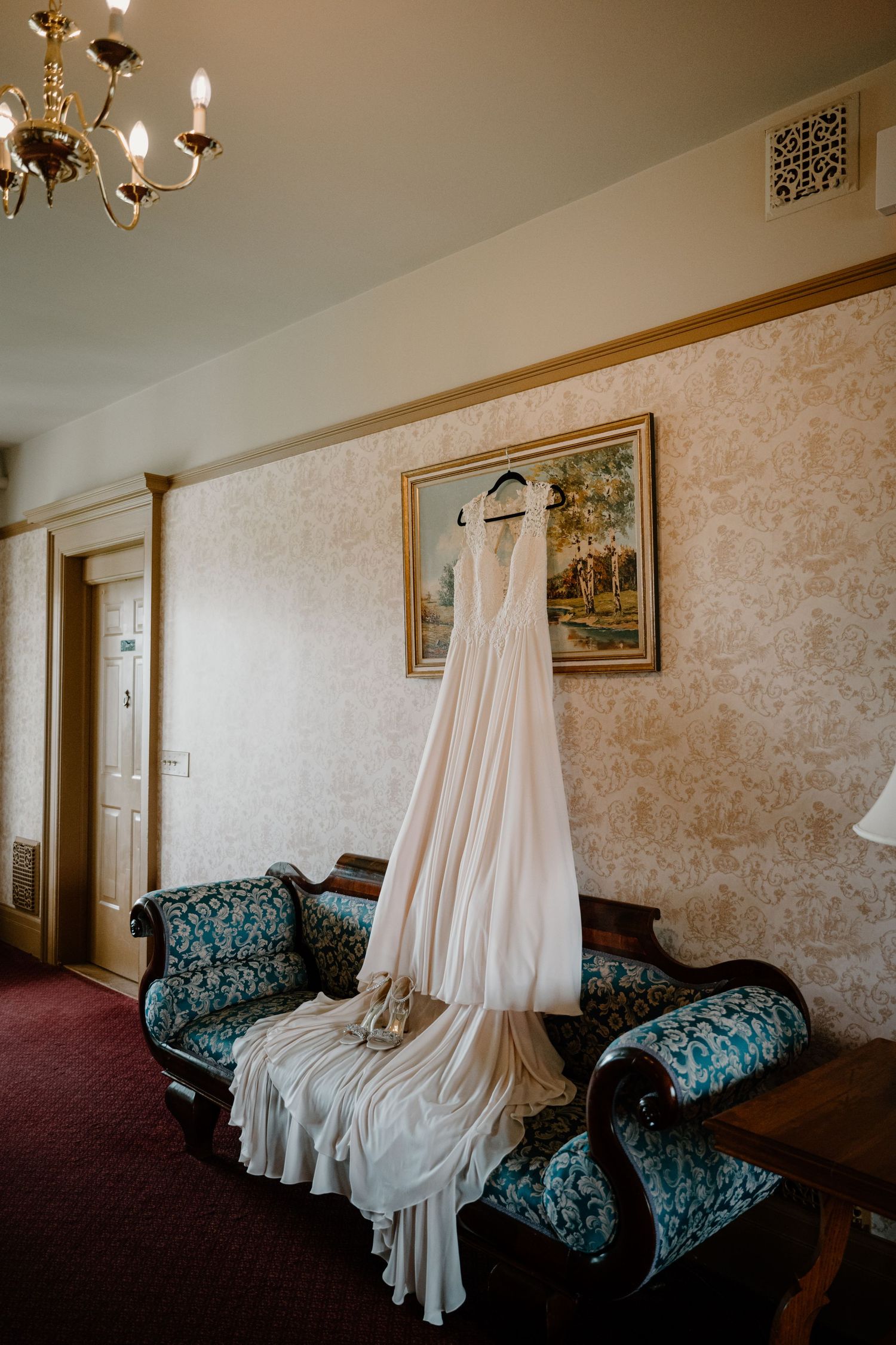 Elegant white wedding gown hanging above vintage teal sofa in ornate wallpapered hotel room with chandelier.