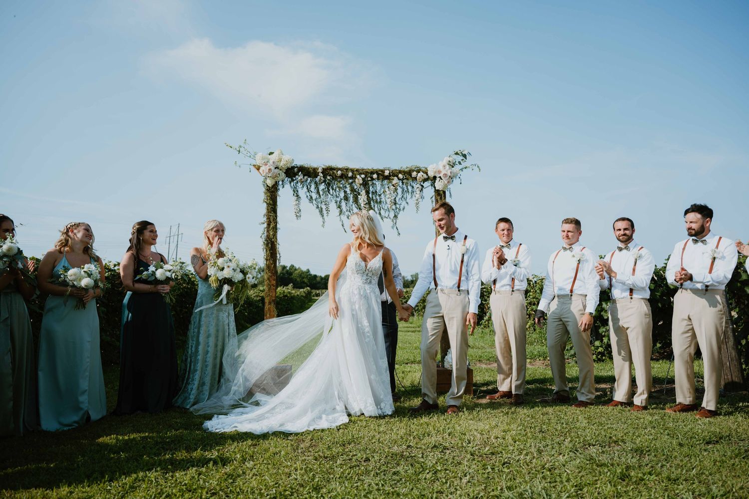 Joyful bride and groom standing at outdoor altar with wedding party under floral arch on sunny day.