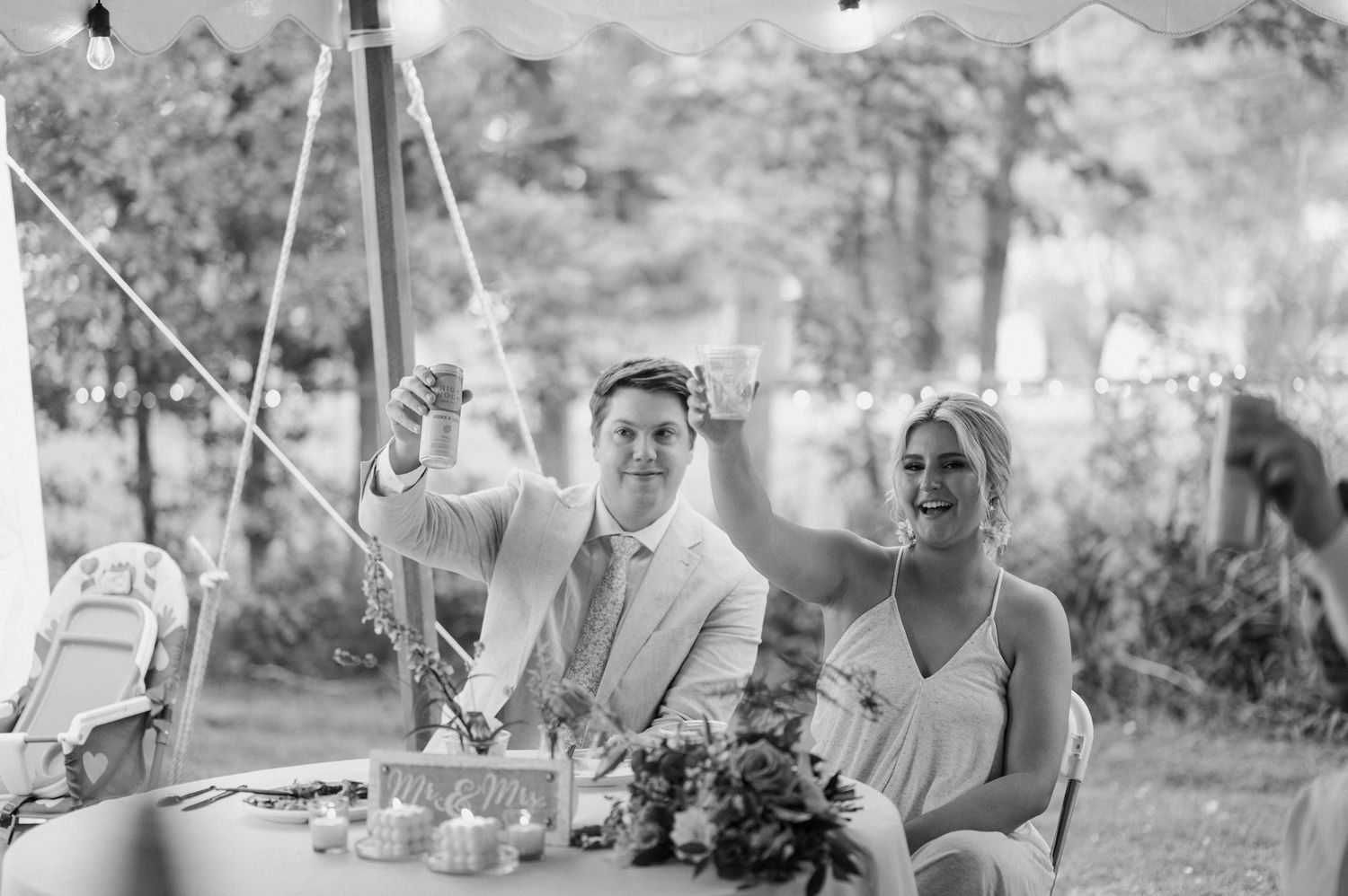 Bride and groom smiling and raising glasses during wedding reception under a white tent.