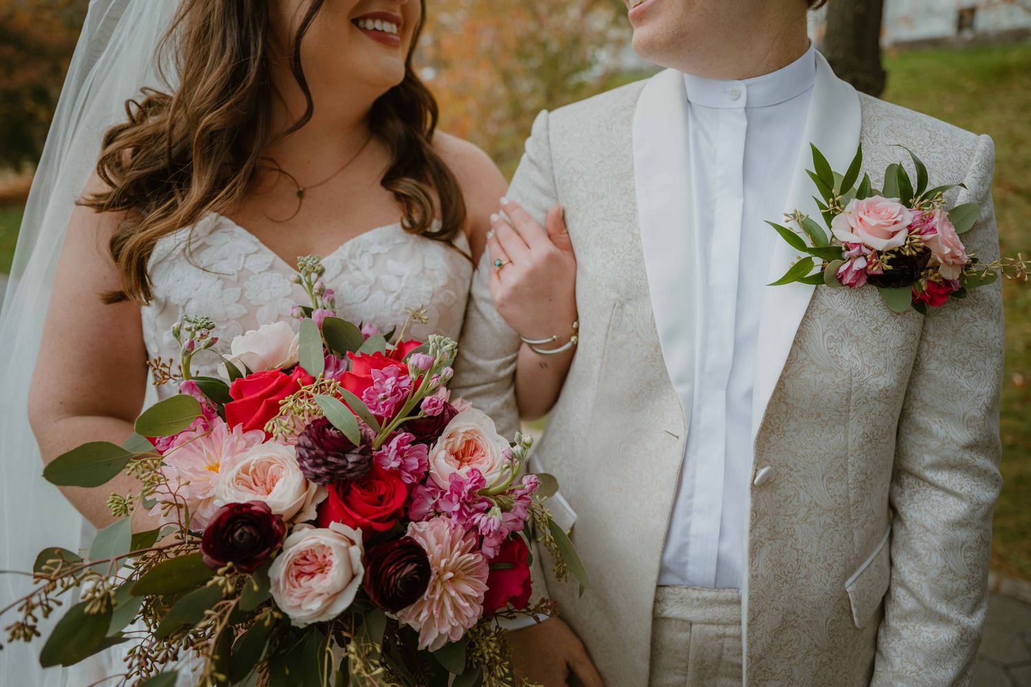 Bride in lace gown holding vibrant pink and red bouquet stands with partner in cream suit at fall wedding.