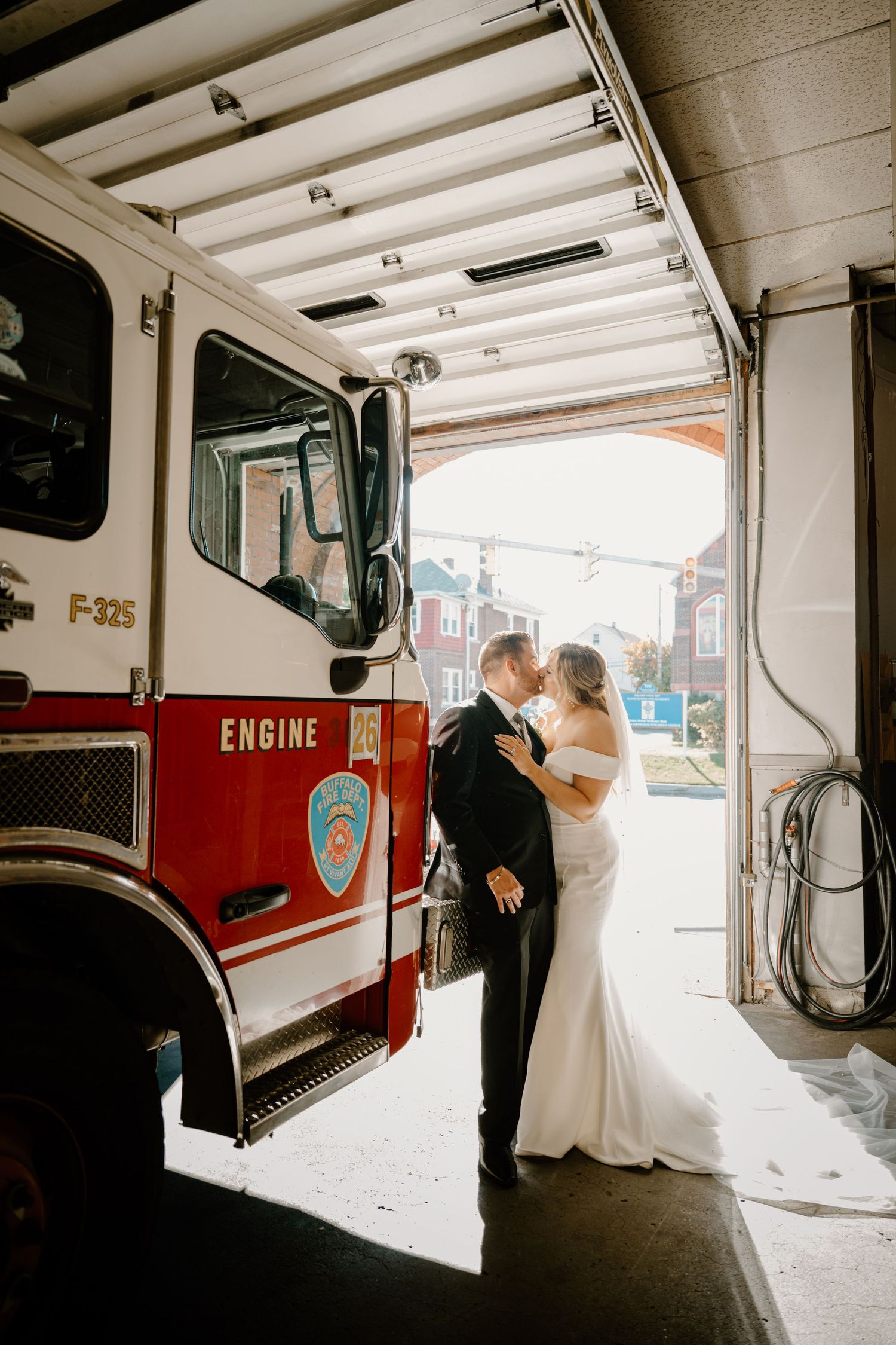 Bride and groom kiss romantically beside a red fire engine inside a sunny firehouse garage.