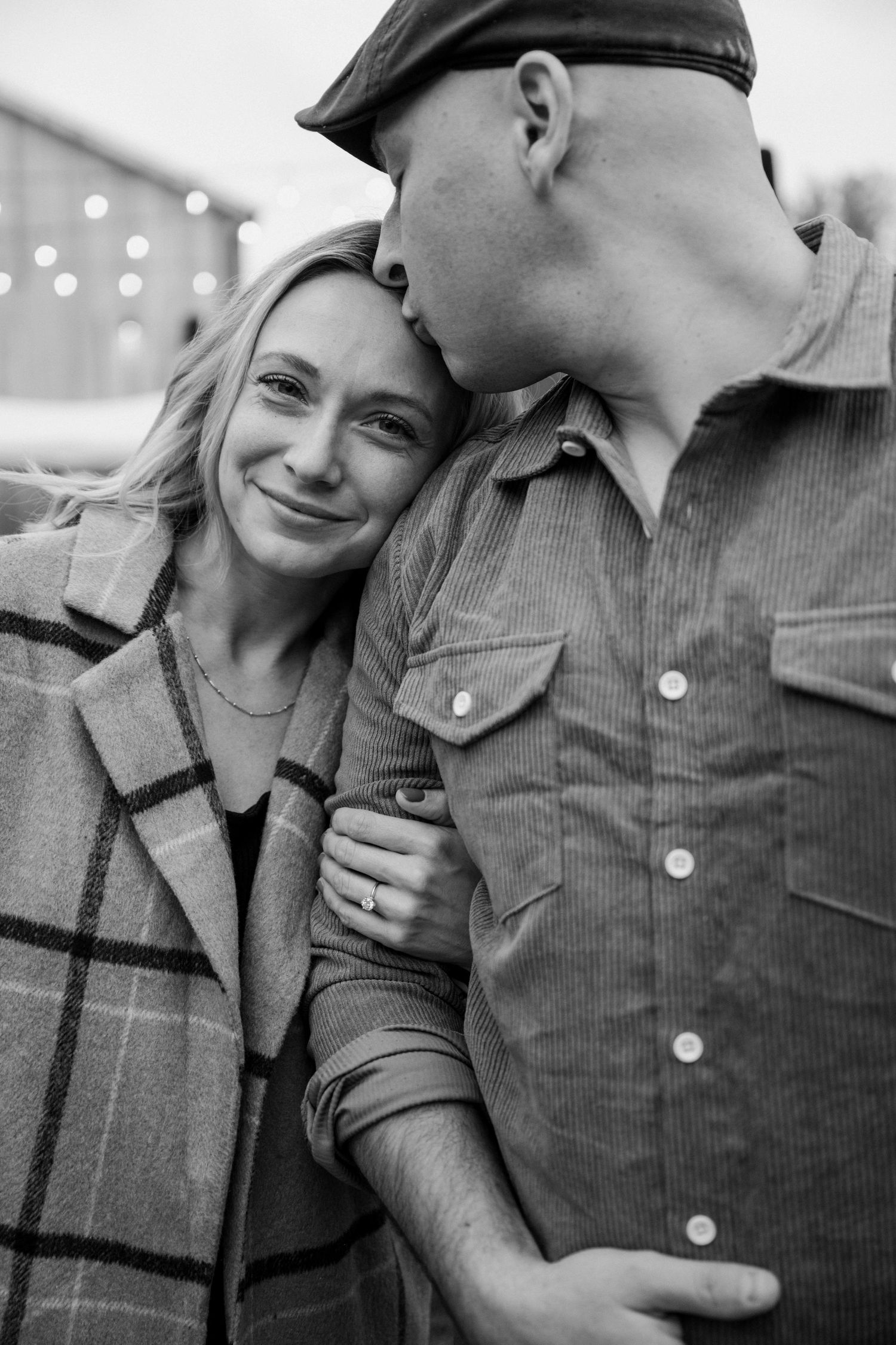 Black and white photo of man kissing woman on forehead as she smiles wearing plaid coat.