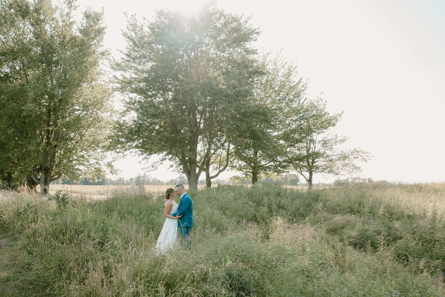Bride and groom walk hand in hand through lush green meadow with tall trees and golden sunlight in background.