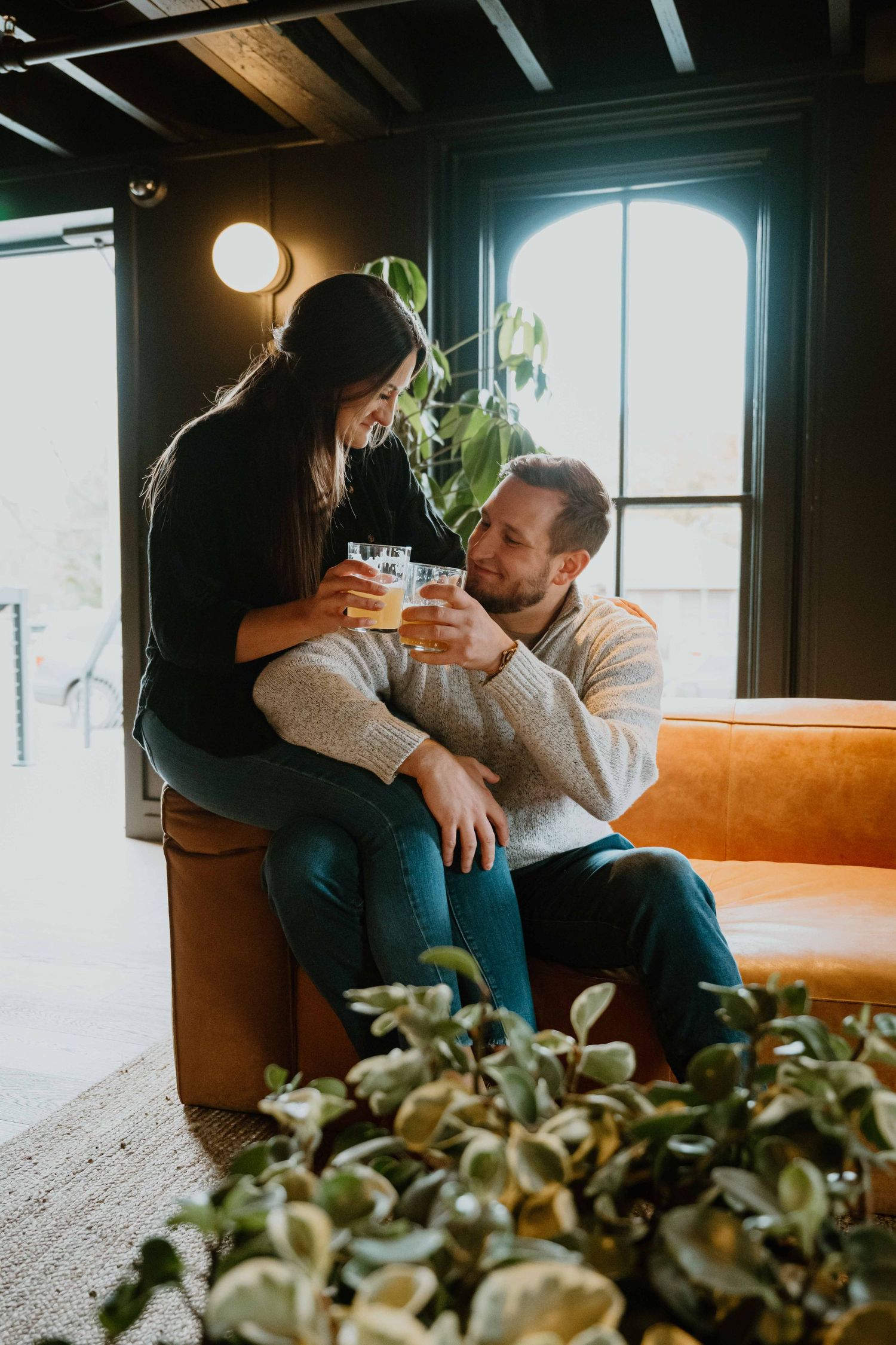 Happy couple toasting with craft beers on orange leather sofa in stylish dark-walled lounge.