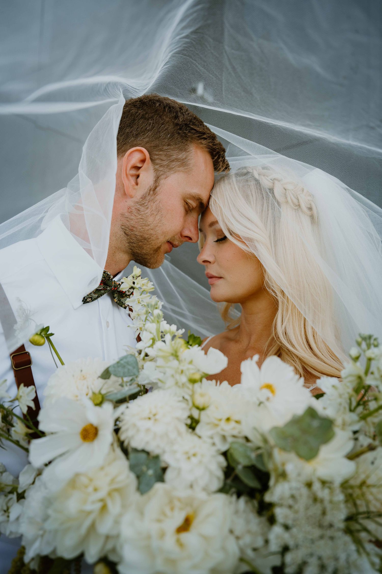Intimate portrait of bride and groom touching foreheads under flowing veil with white floral bouquet.