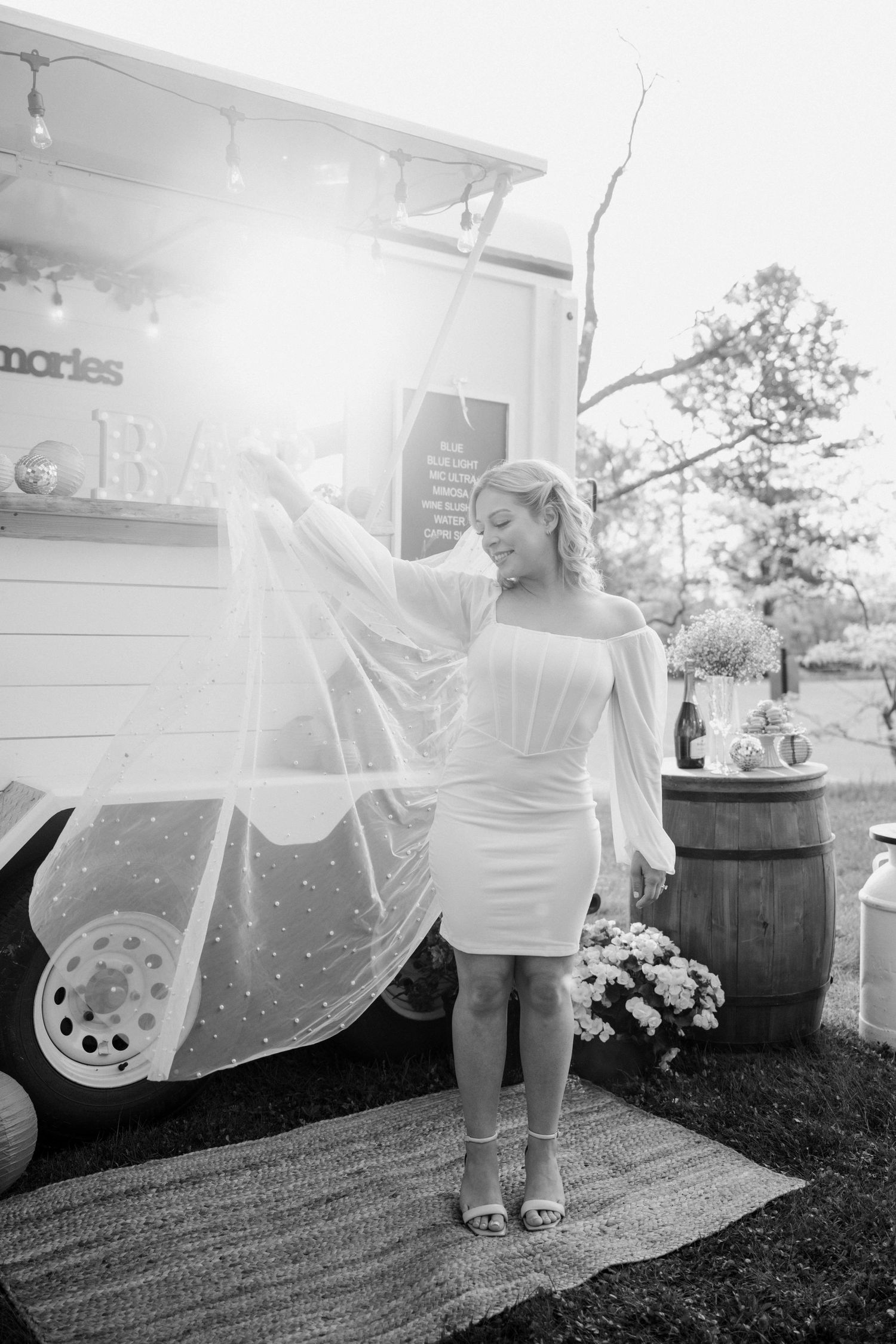 Black and white photo of bride in short dress twirling veil near a vintage bar trailer.