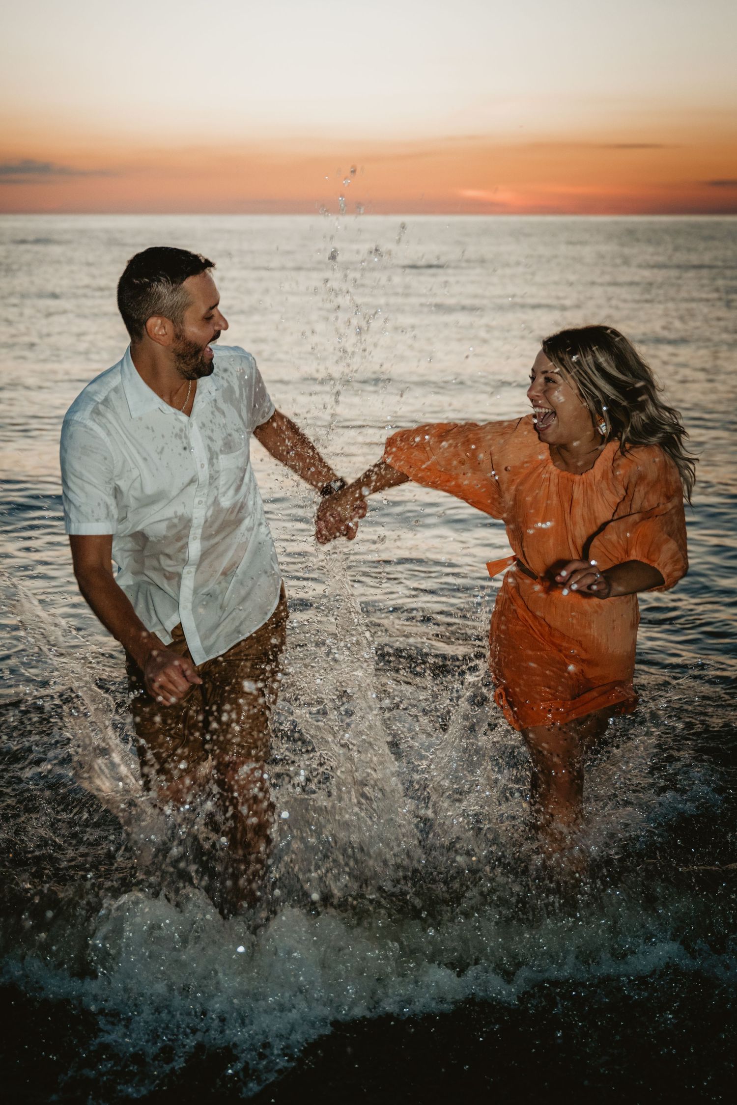 Joyful couple splashing in ocean waves at sunset, holding hands in wet clothing.