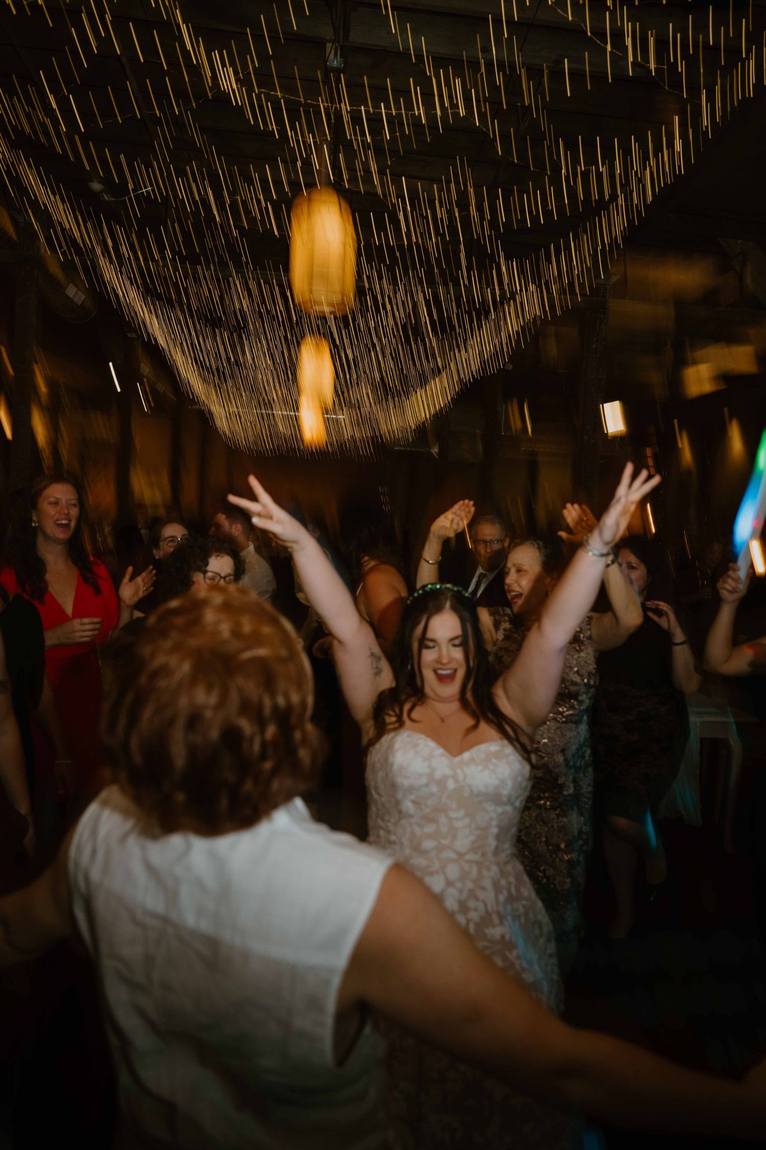 Joyful bride in lace gown raises arms while dancing at a festive wedding reception with guests.