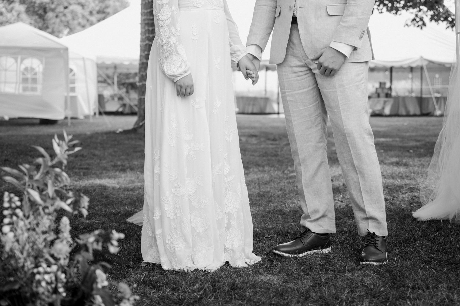 Black and white photo of bride and groom holding hands in wedding attire on a lawn.