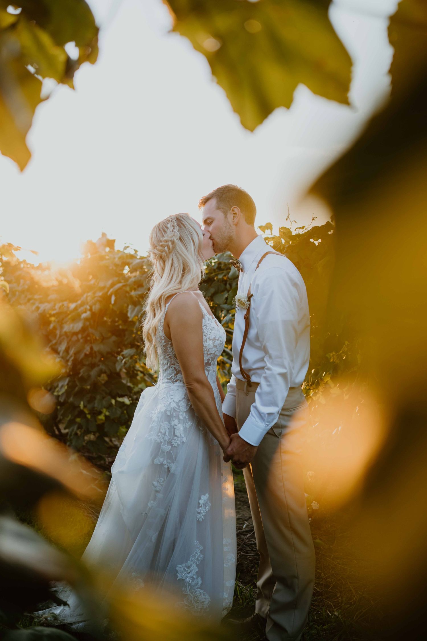 Bride and groom share a golden hour kiss framed by vineyard leaves in warm sunset light.