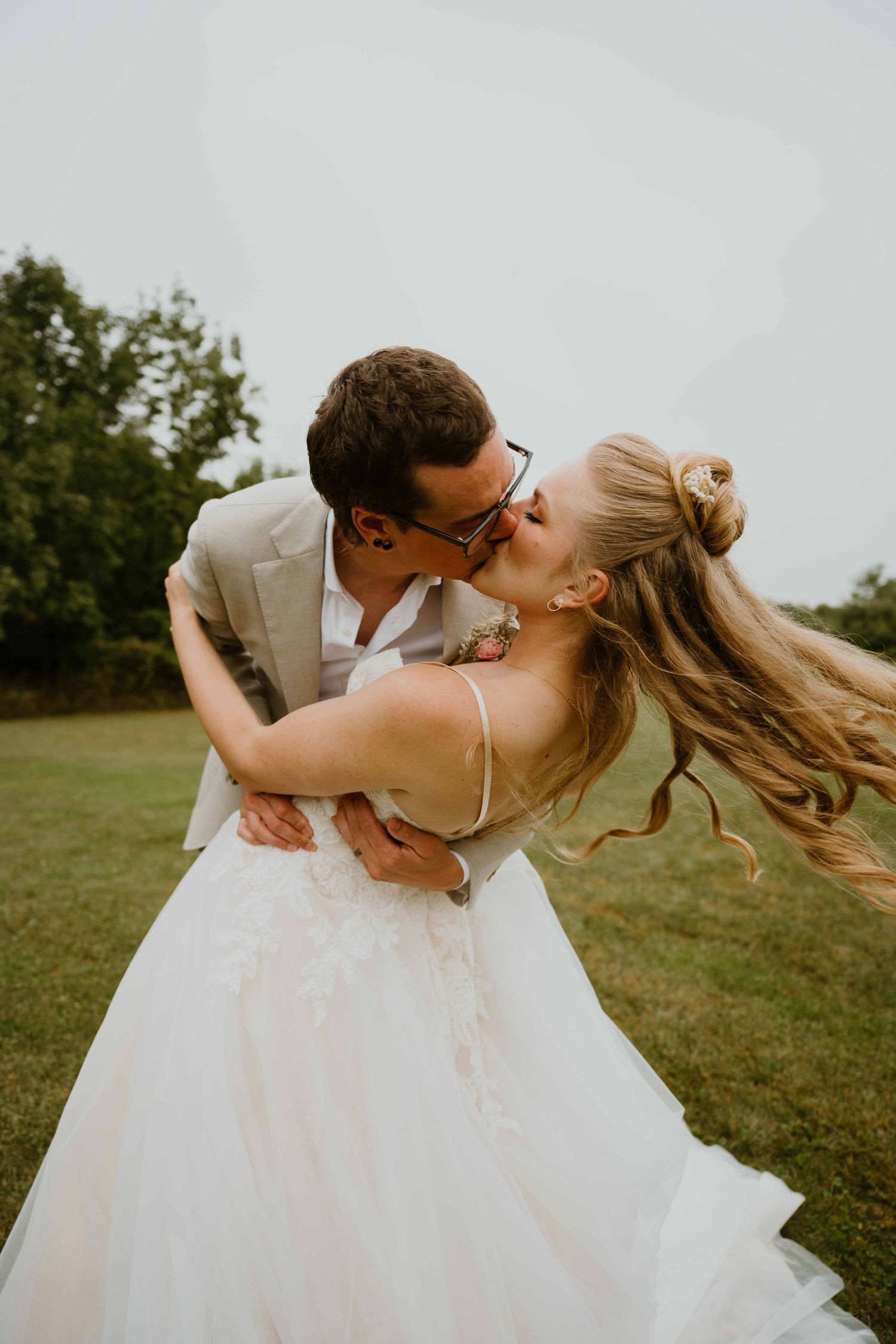 Groom dips bride for a kiss in field, her hair flowing, both in wedding attire.