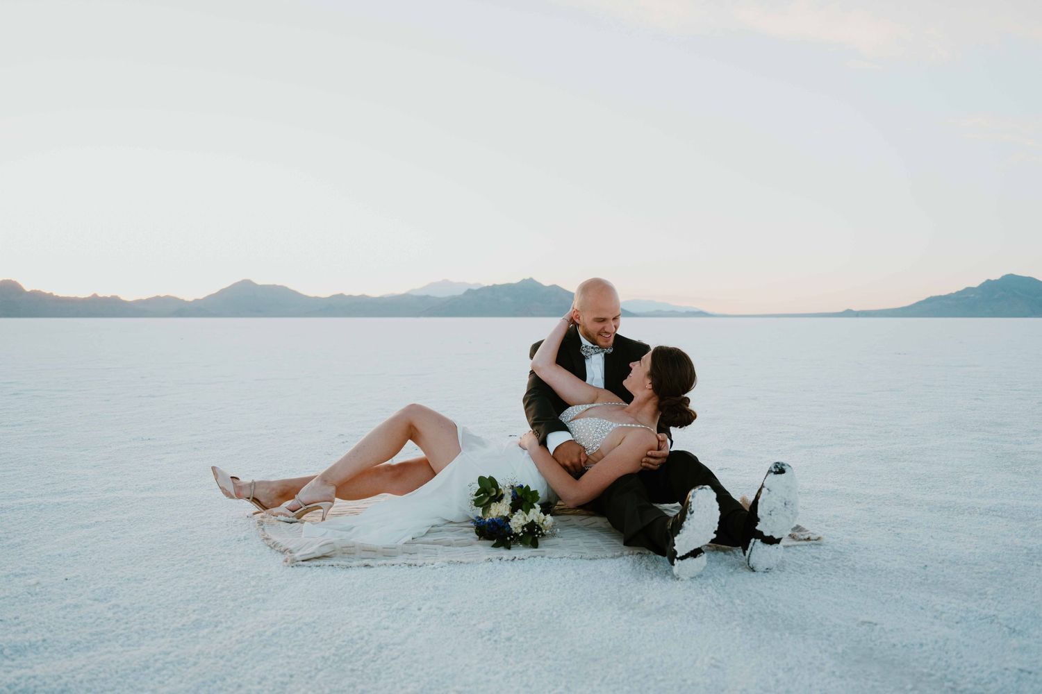 Bride and groom sit intimately together on the white salt flats with mountains behind them.