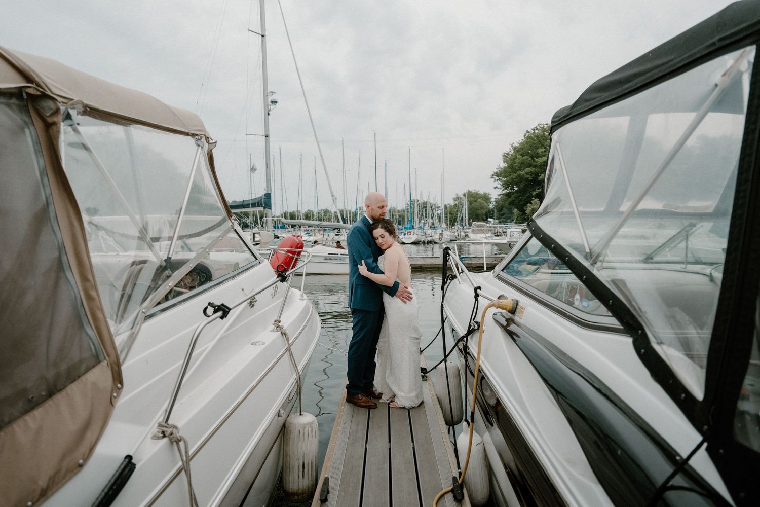 Bride and groom share romantic embrace between sailboats at marina dock on their wedding day.