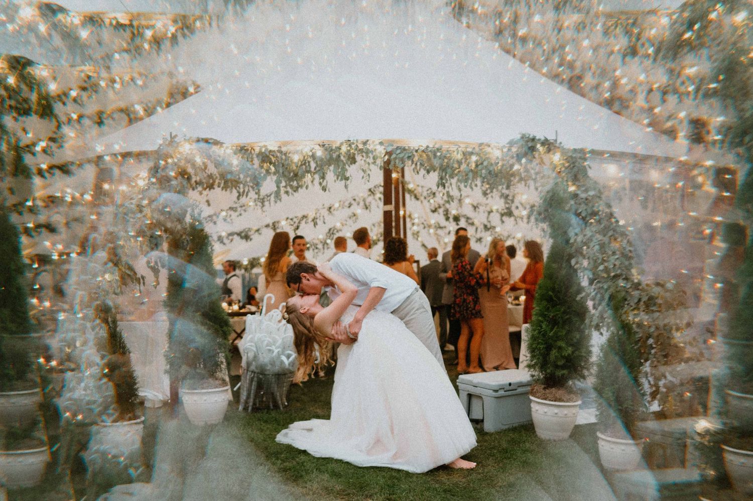 Bride and groom share romantic dip kiss under glowing tent surrounded by wedding guests.
