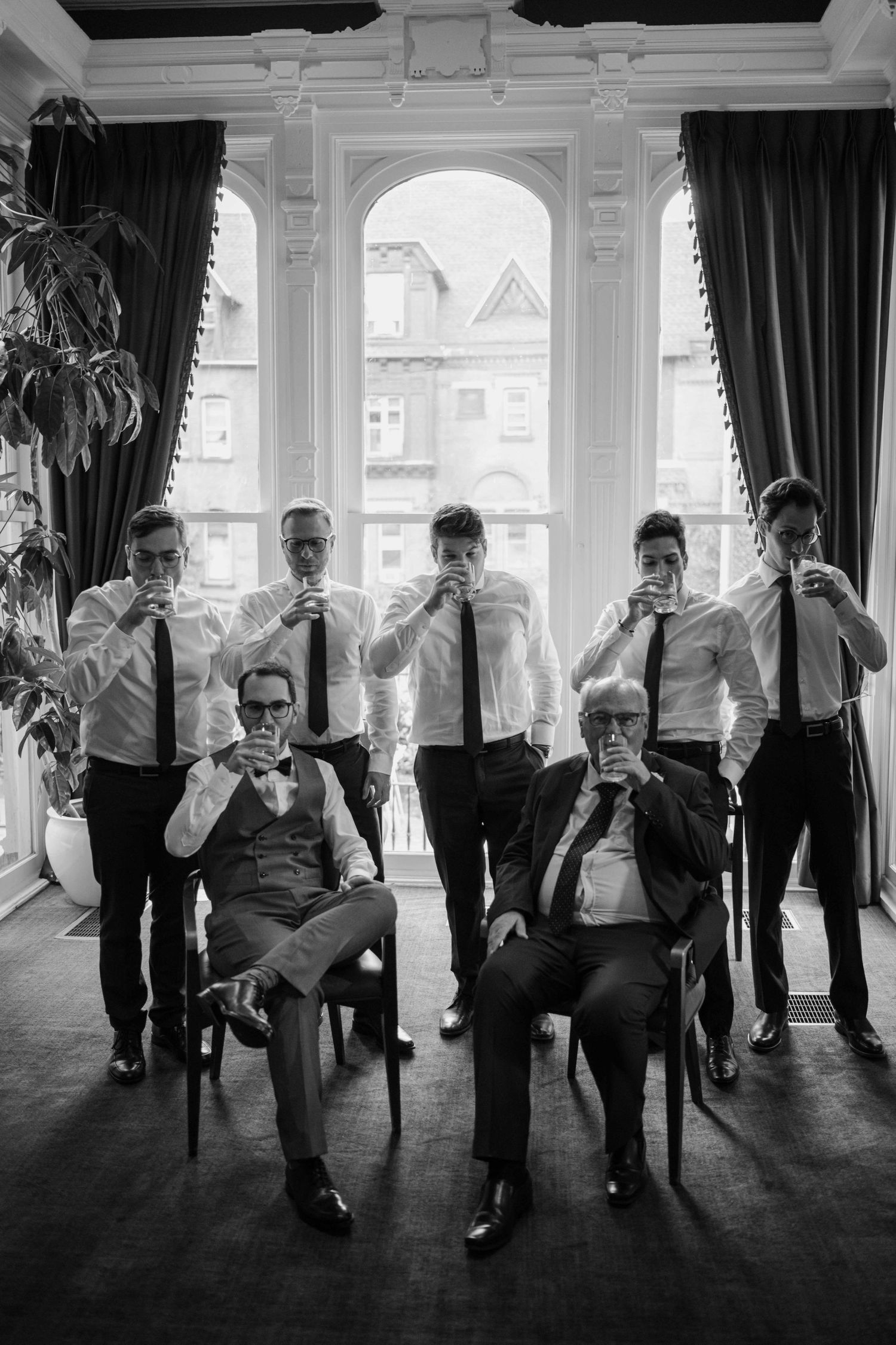 Groom and groomsmen in black and white photo toasting with drinks near large arched windows.