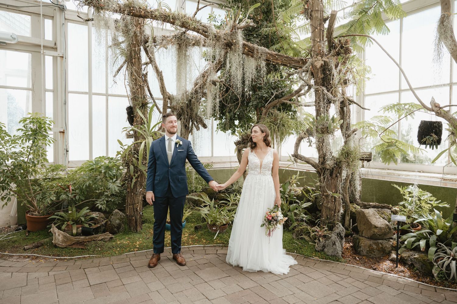 Bride and groom holding hands in botanical greenhouse surrounded by tropical plants.