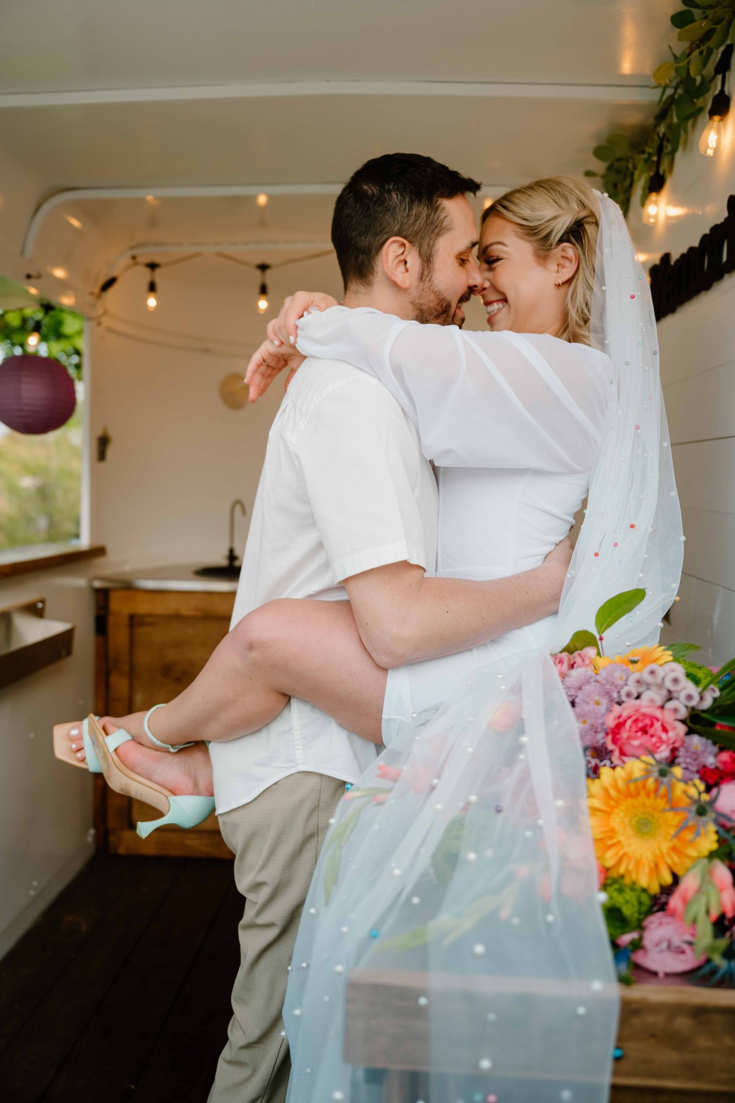 Groom lifts smiling bride in white mini dress and veil inside a decorated mobile bar truck.