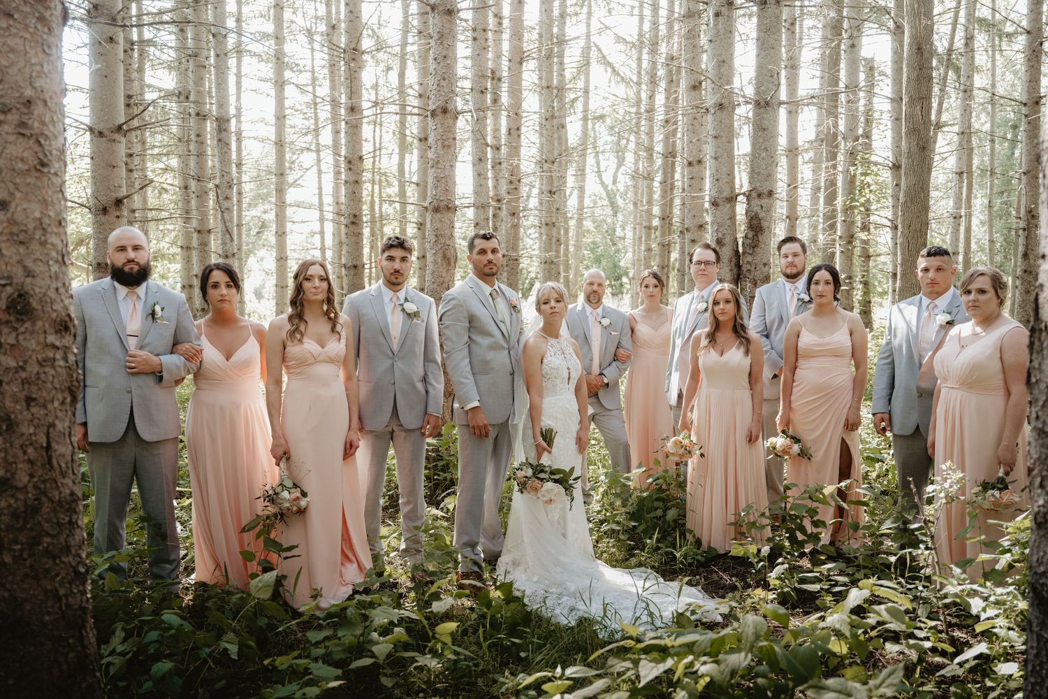 Wedding party poses in peach and gray attire among tall birch trees in enchanting forest setting.