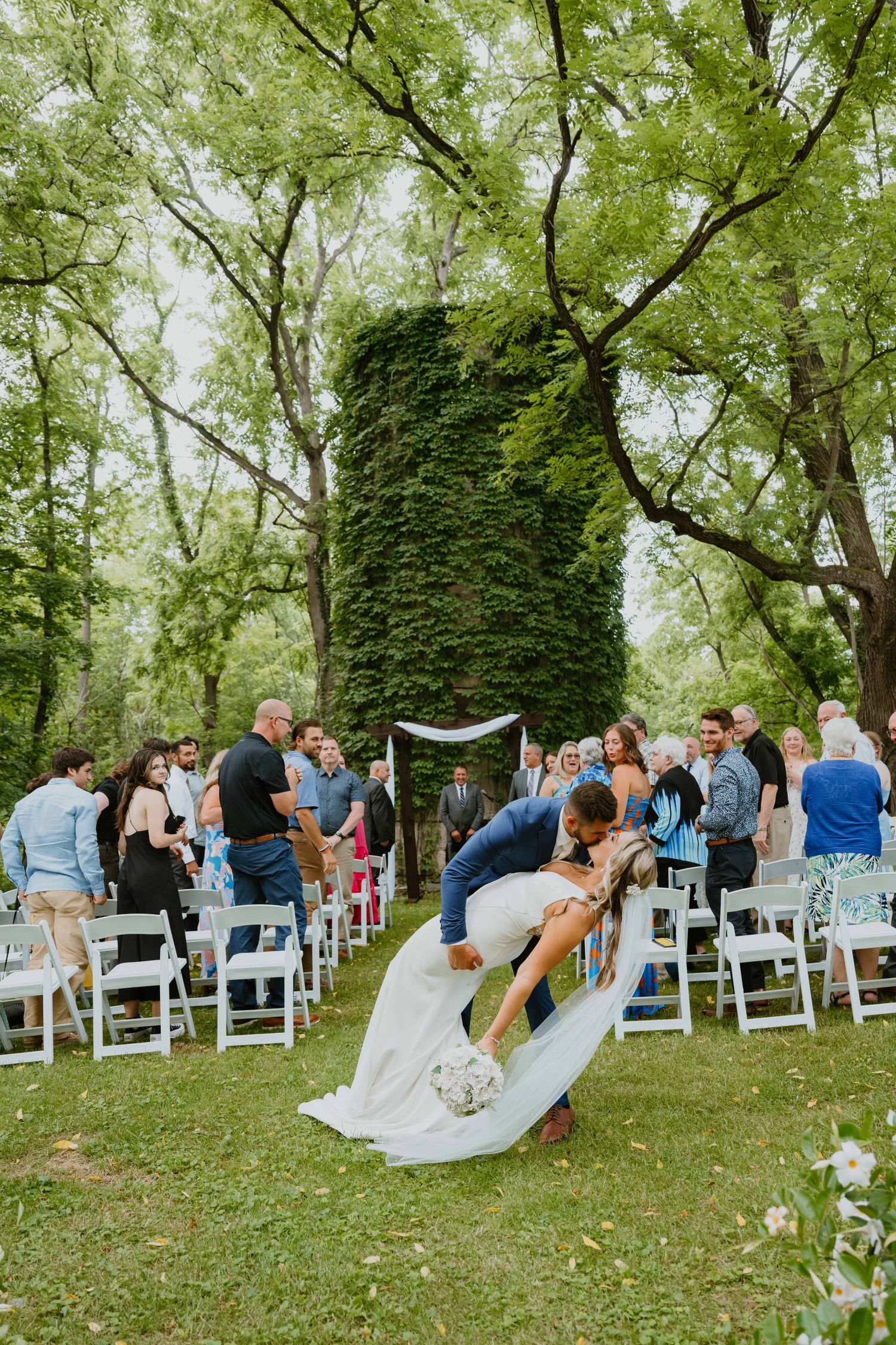 Groom dips bride for romantic kiss during outdoor wedding recessional with ivy tower backdrop.