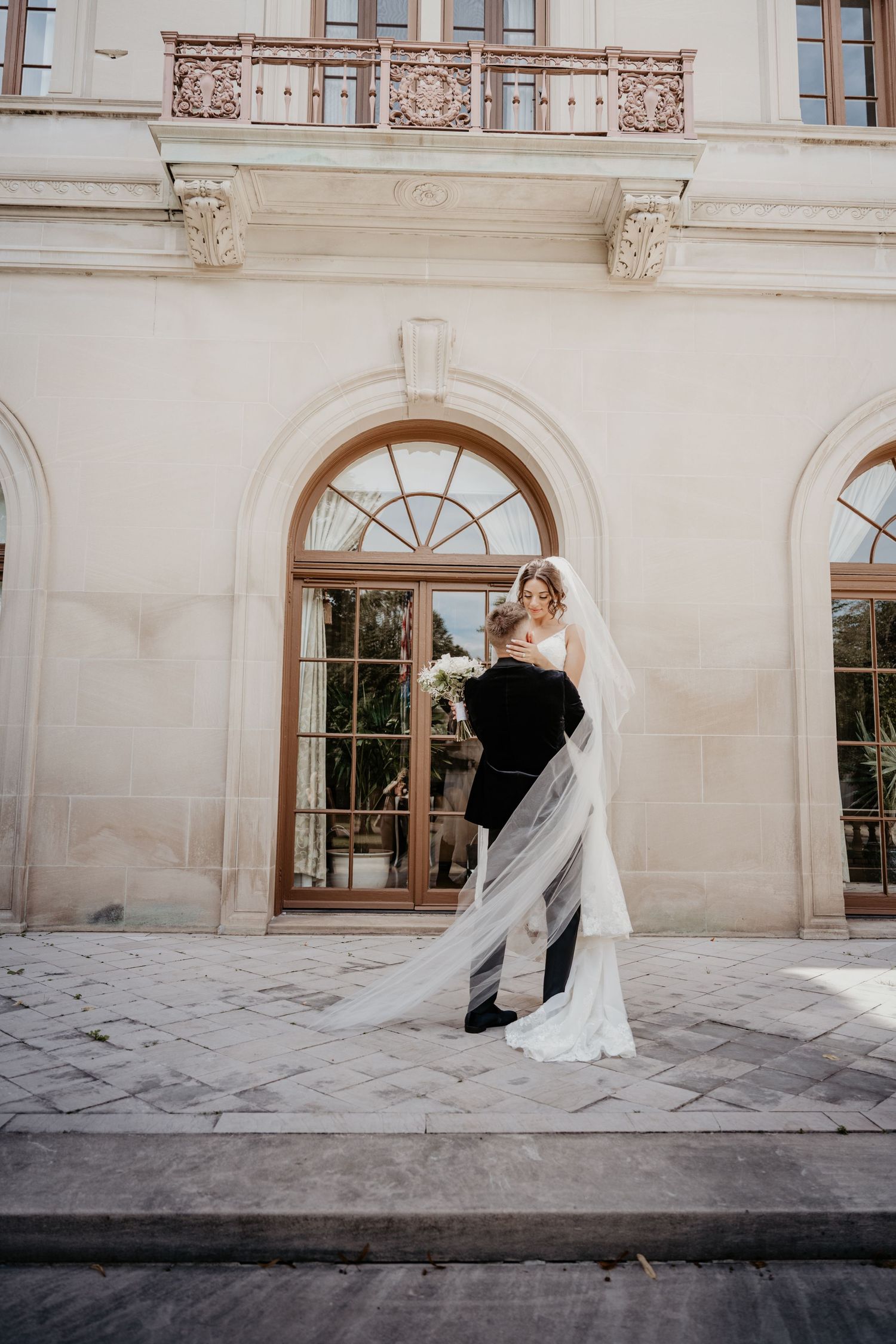 Groom lifting bride in white gown and cathedral veil outside ornate classical building.