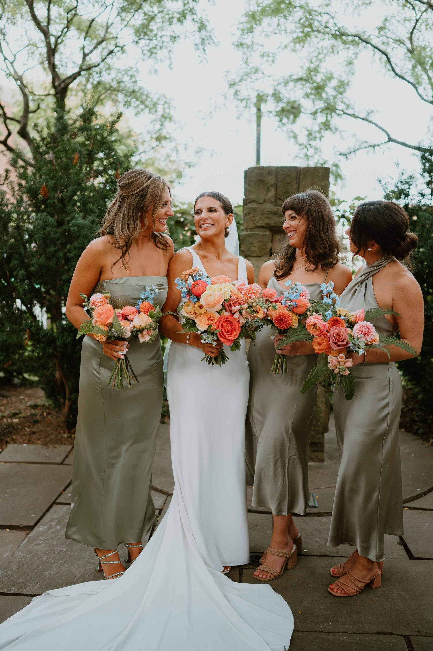 Bride in white gown laughs with three bridesmaids in sage green satin dresses holding colorful floral bouquets outdoors.