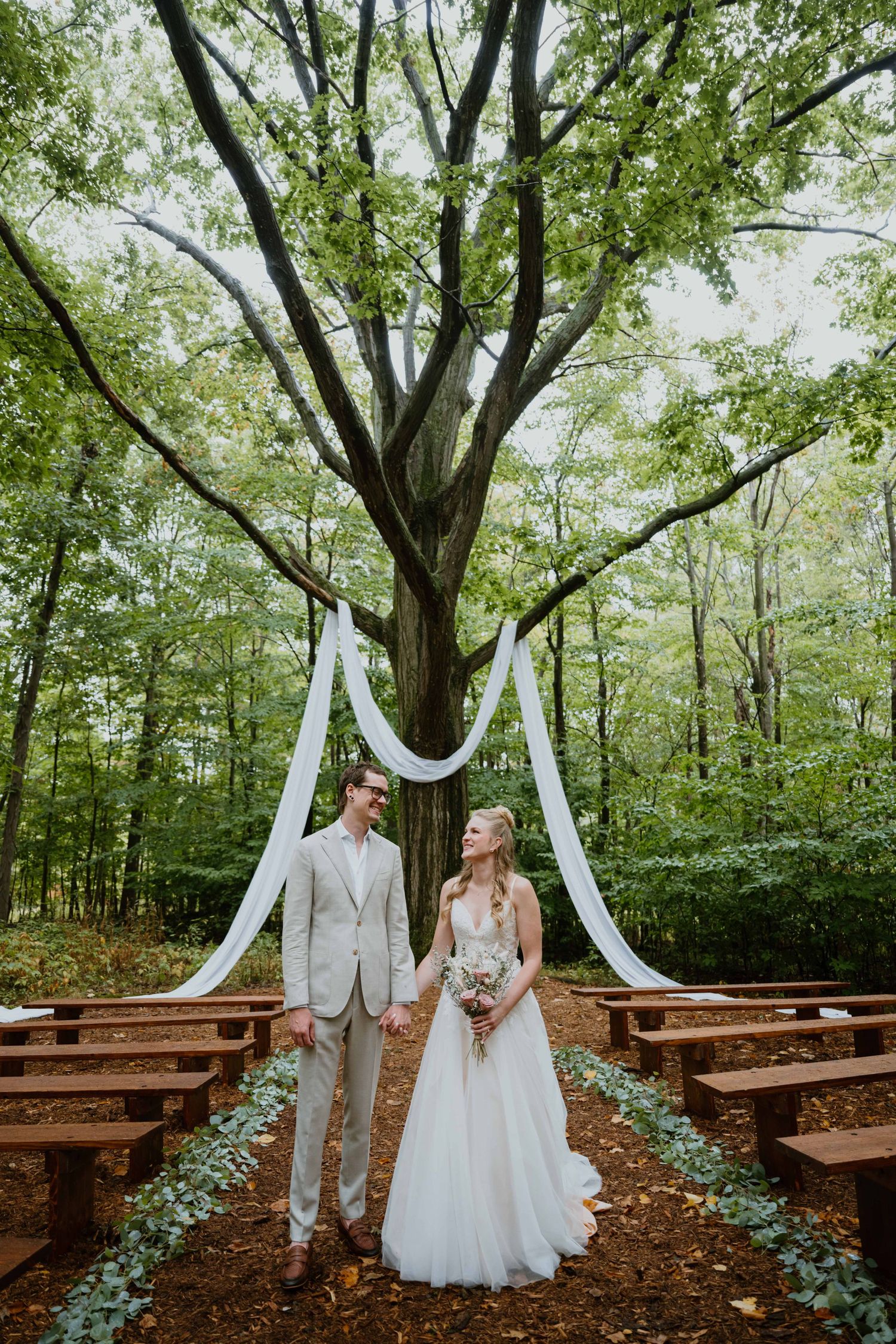 Bride and groom hold hands beneath a large tree with white draping at forest ceremony.