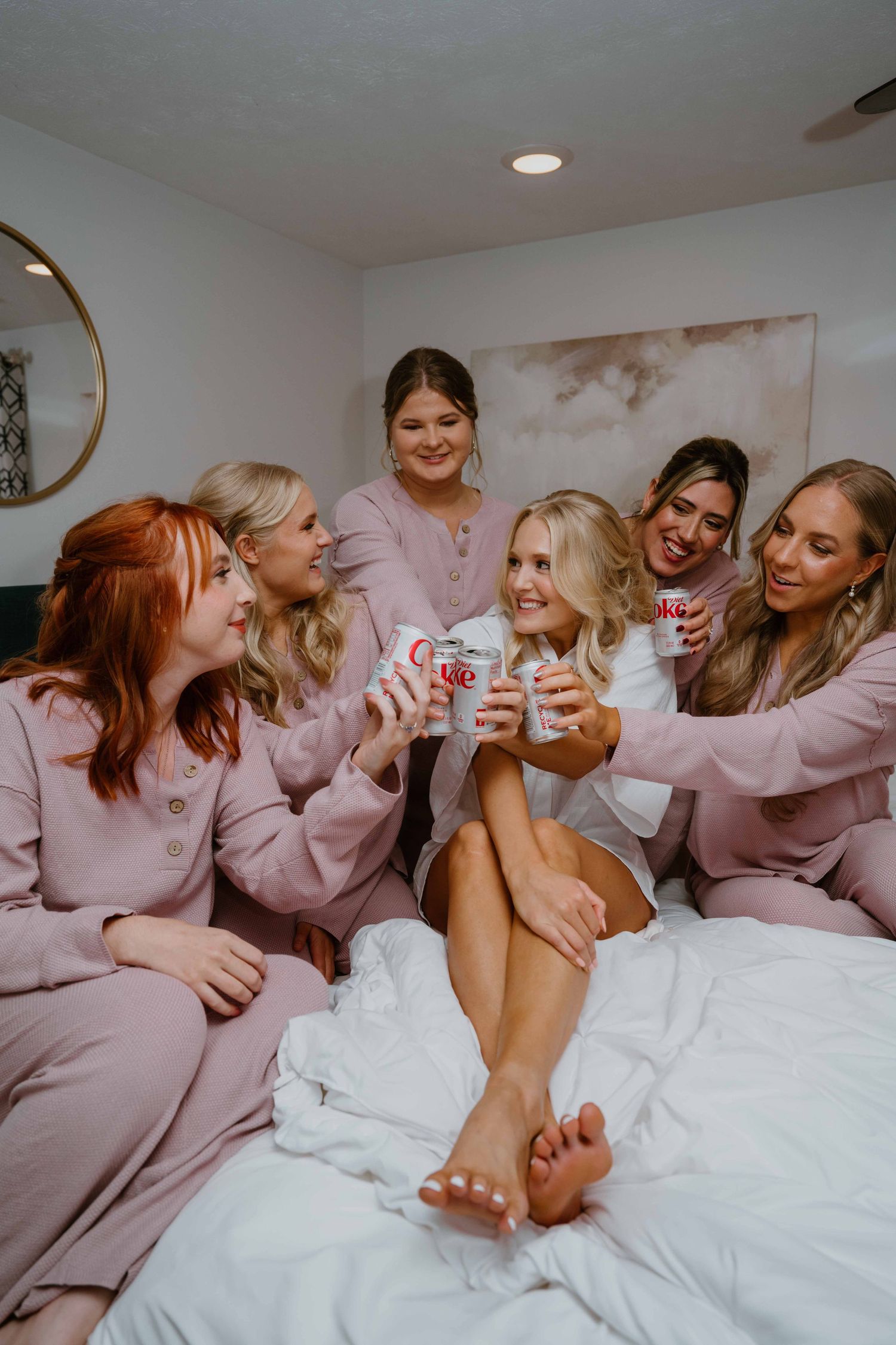 Bride celebrating with bridesmaids in pink pajamas toasting with soda cans on white bed.
