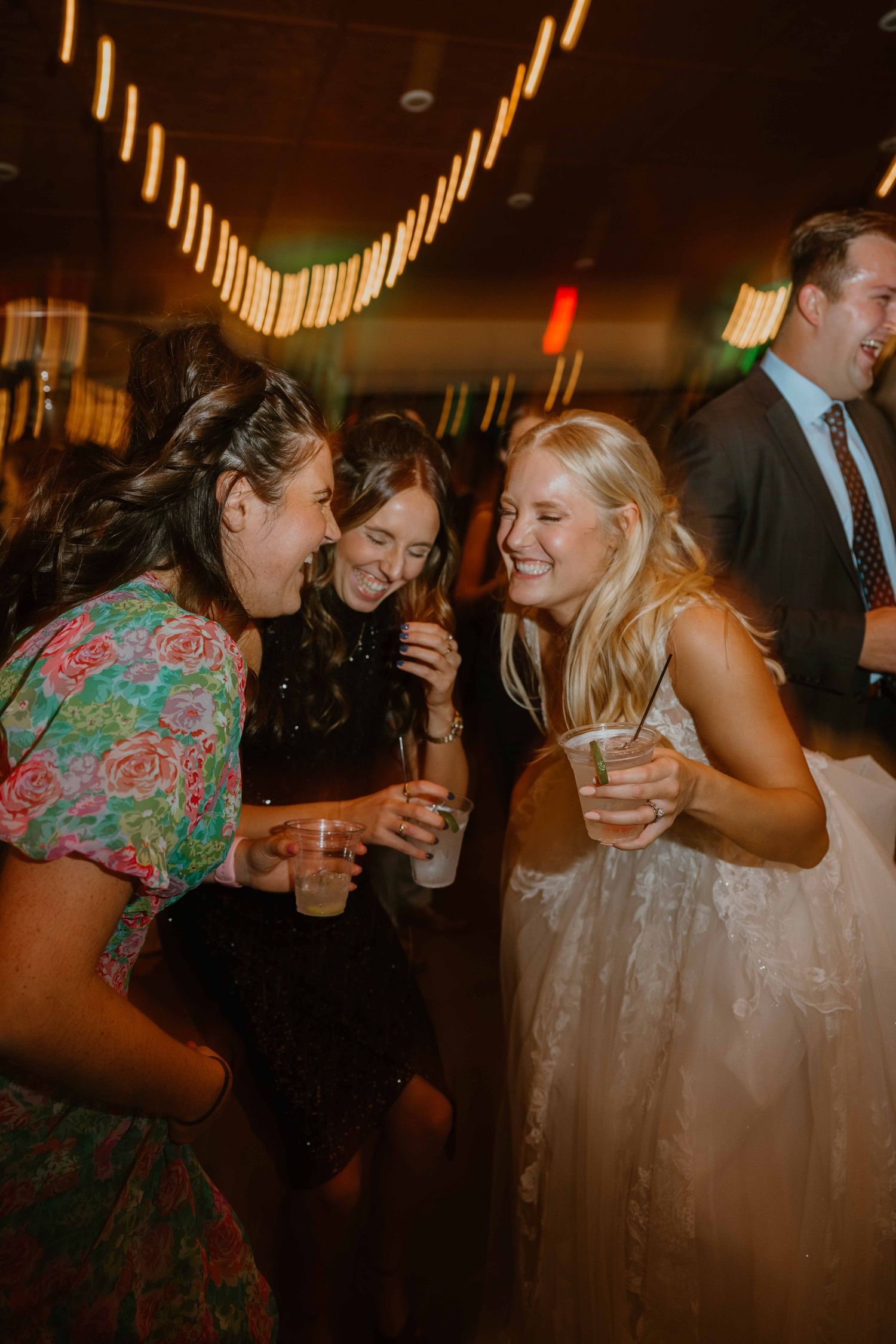 Three women laughing and holding drinks at a wedding reception with string lights.