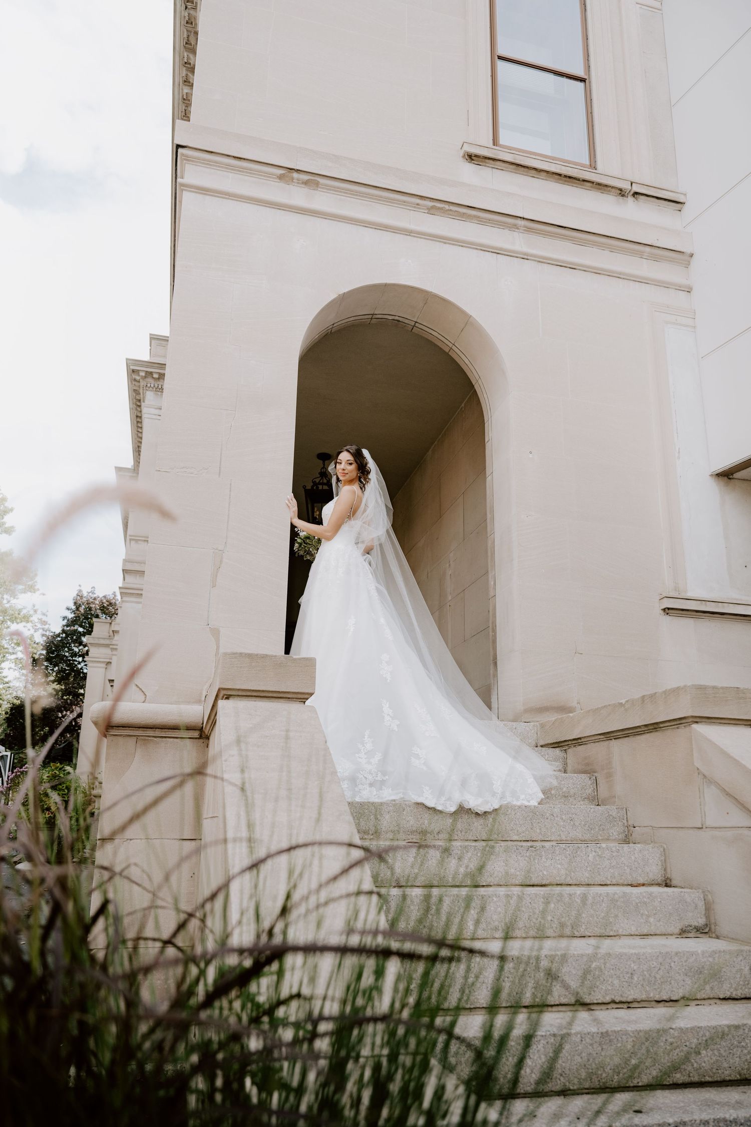 Bride in flowing white gown and veil descending stone steps of elegant classical building.